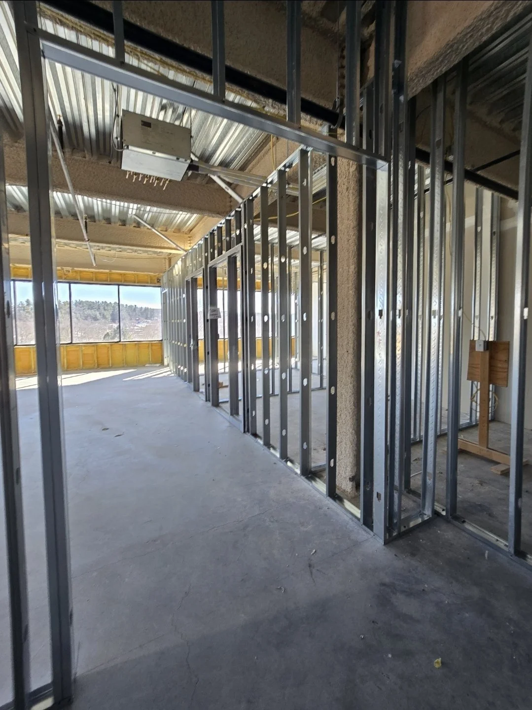 Interior view of a building under construction with metal wall framing and electrical wiring, large windows showing a landscape outside, and construction materials on the floor.