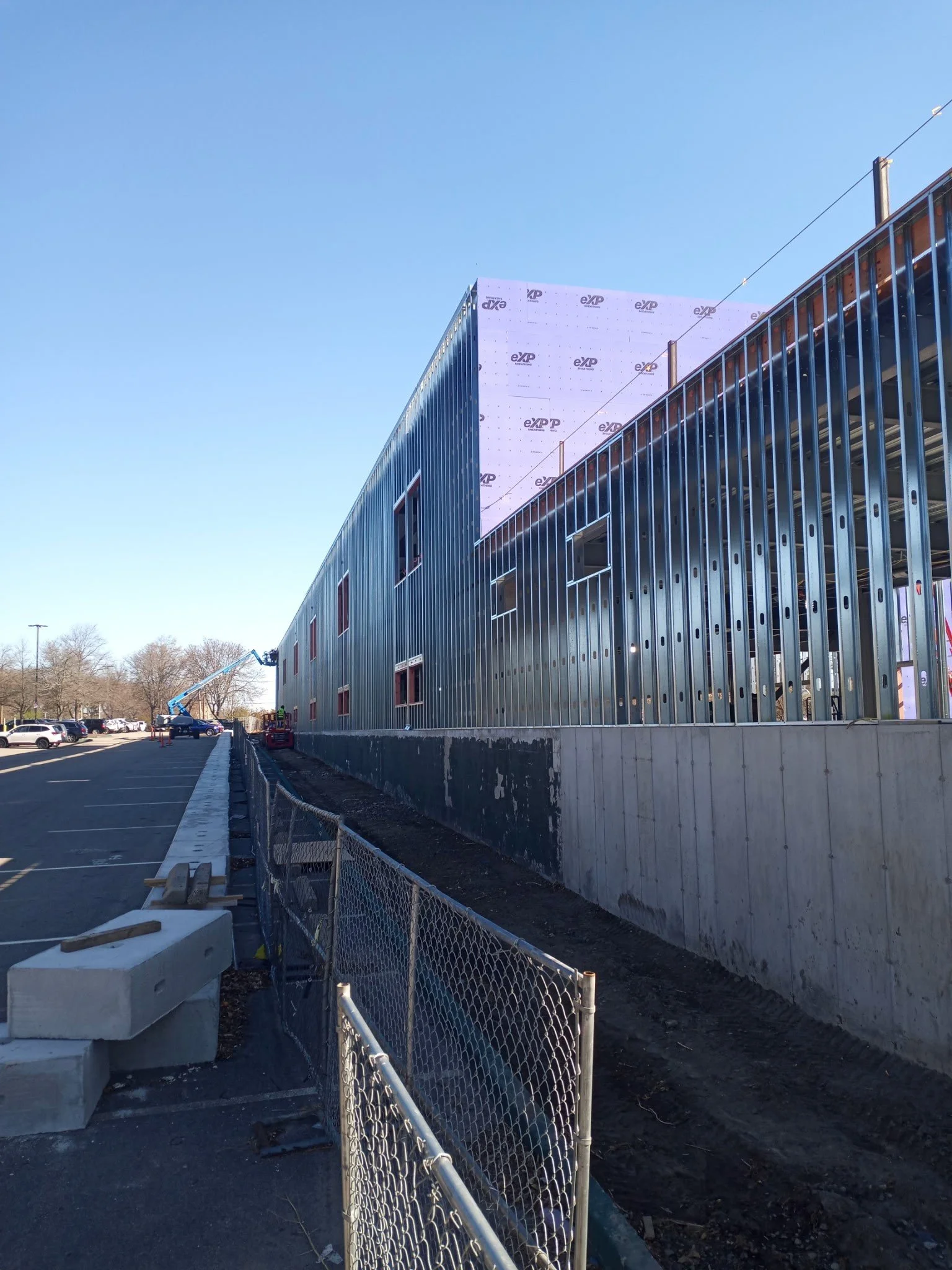 Construction site with an unfinished building featuring metal framing and insulation, a construction lift, concrete barriers, and a parking lot with parked cars under a clear blue sky.