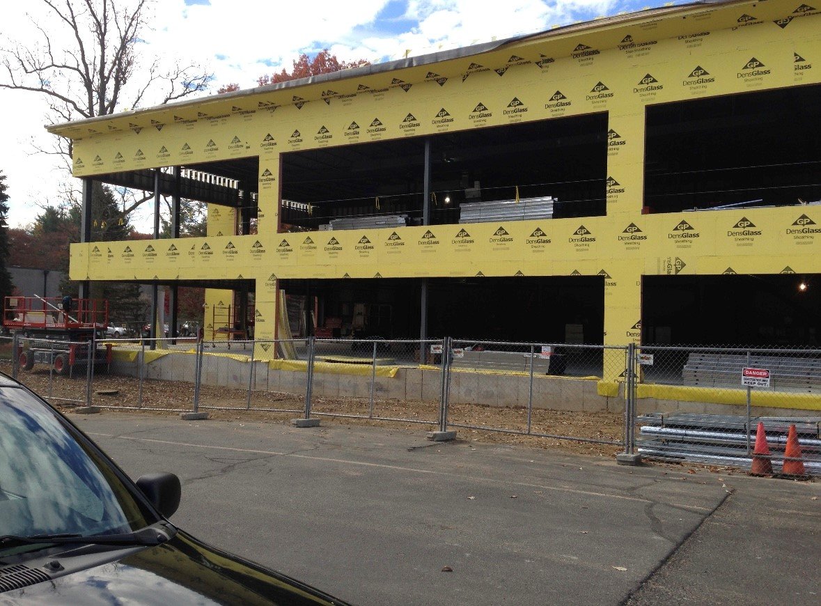 Construction site of a two-story building with yellow sheathing, surrounded by a chain-link fence and orange traffic cones; a parked car is partially visible in the foreground.
