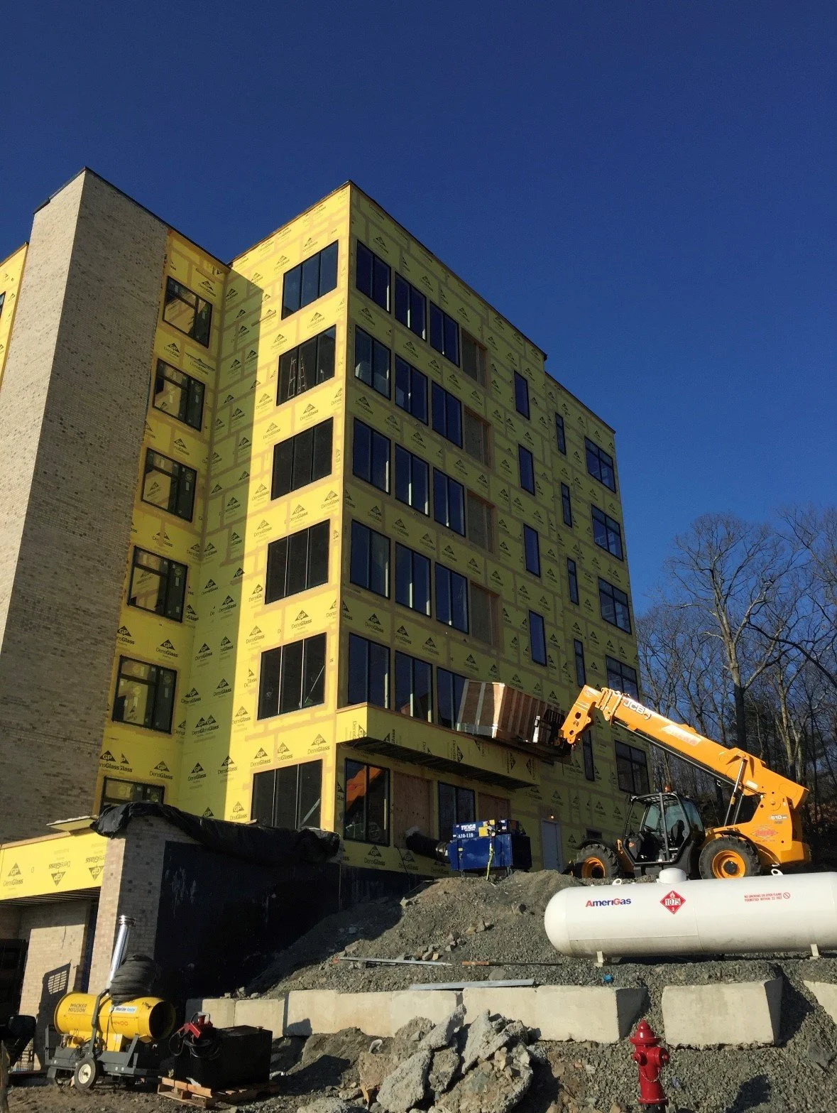 Construction site with a multi-story building under construction, yellow exterior insulation, large windows, construction equipment including a crane, and a clear blue sky.