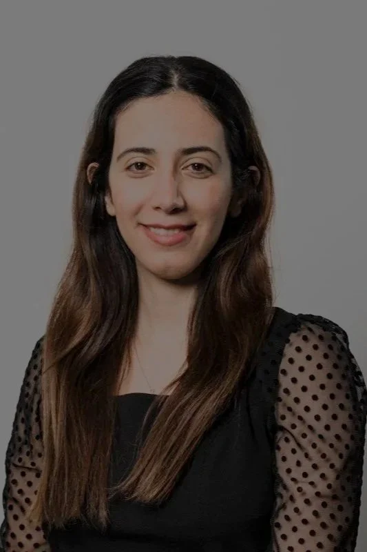 A young woman with long brown hair, smiling, wearing a black top with sheer polka dot sleeves, against a neutral gray background.