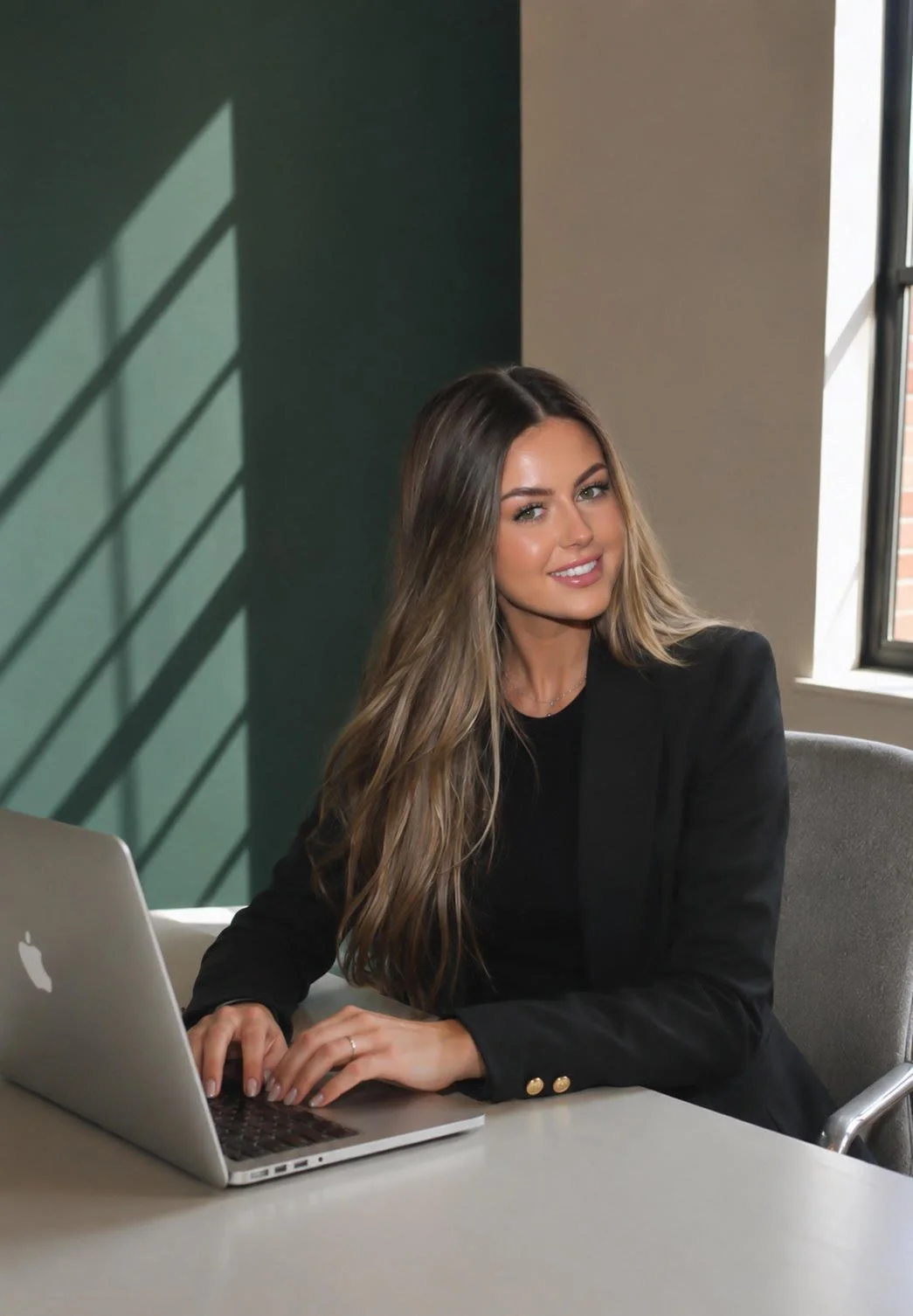 A woman with long wavy hair sitting at a desk with a silver MacBook laptop, wearing a black blazer and smiling at the camera, in a well-lit office space.
