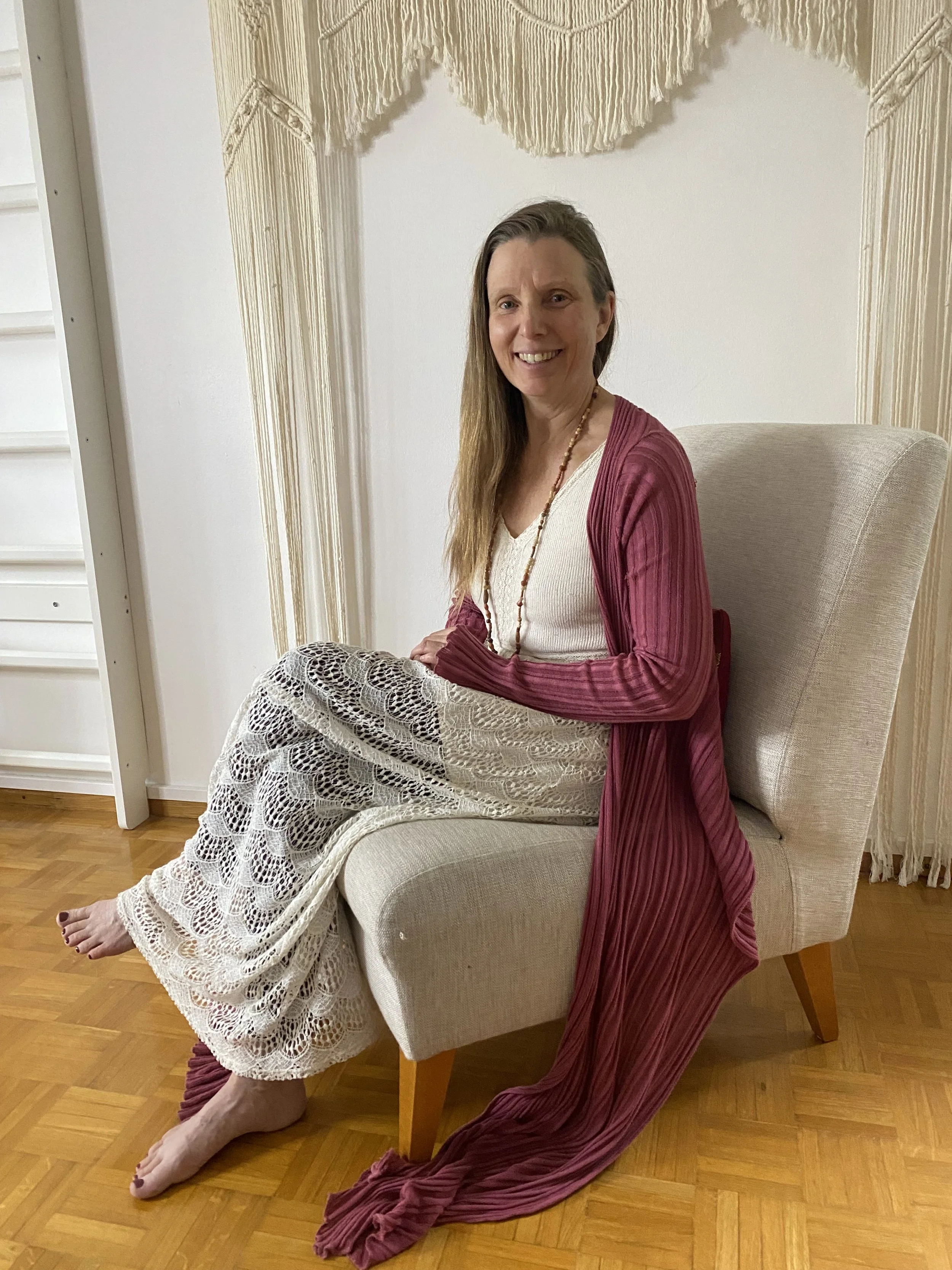 A woman with long hair sitting on a beige armchair in a room with white walls and hardwood floor, wearing a white top, a pink cardigan, and a long skirt, smiling at the camera.