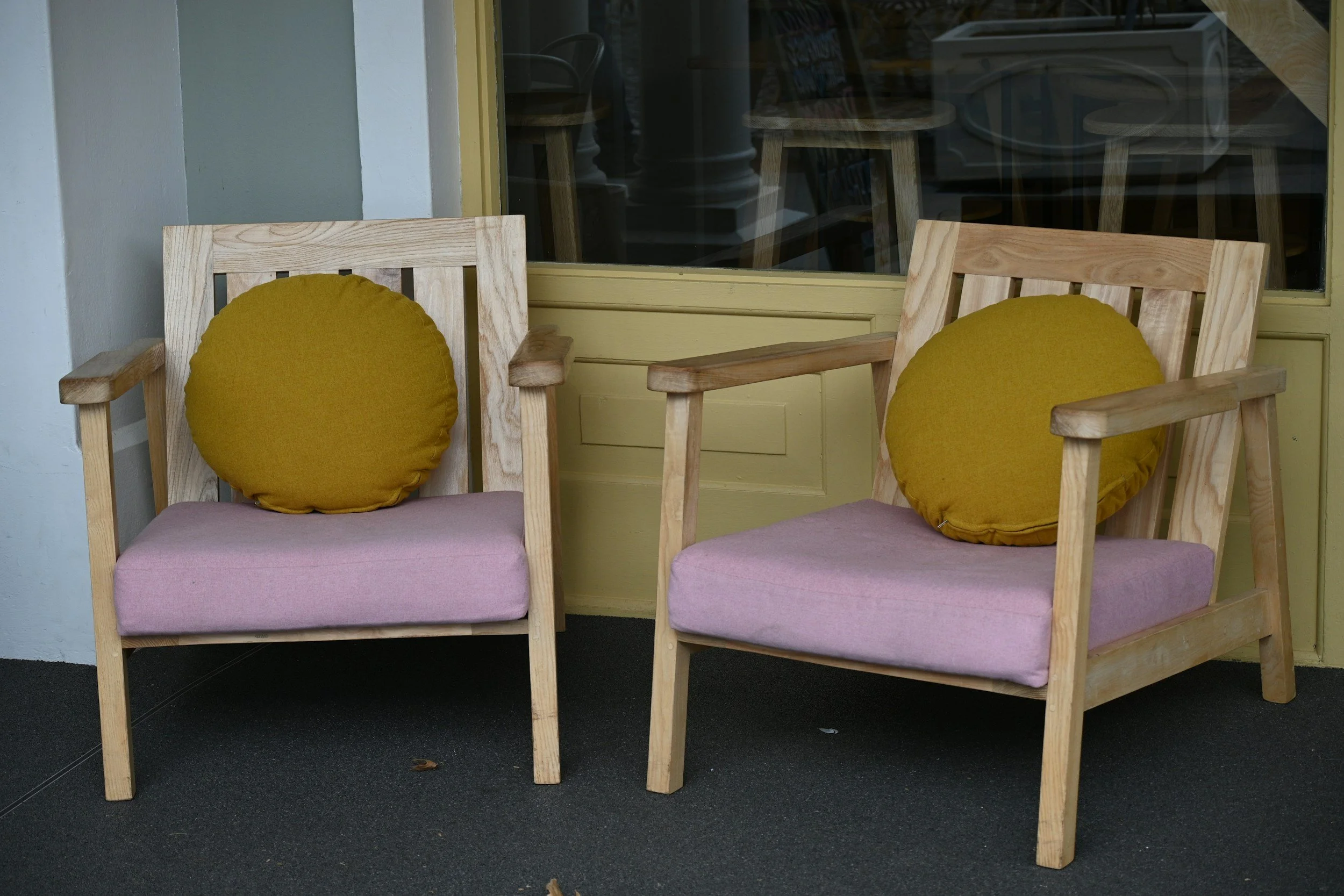 Two wooden chairs with yellow cushions and pink seat pads are placed side by side in front of a window with a yellow frame, in an indoor setting.