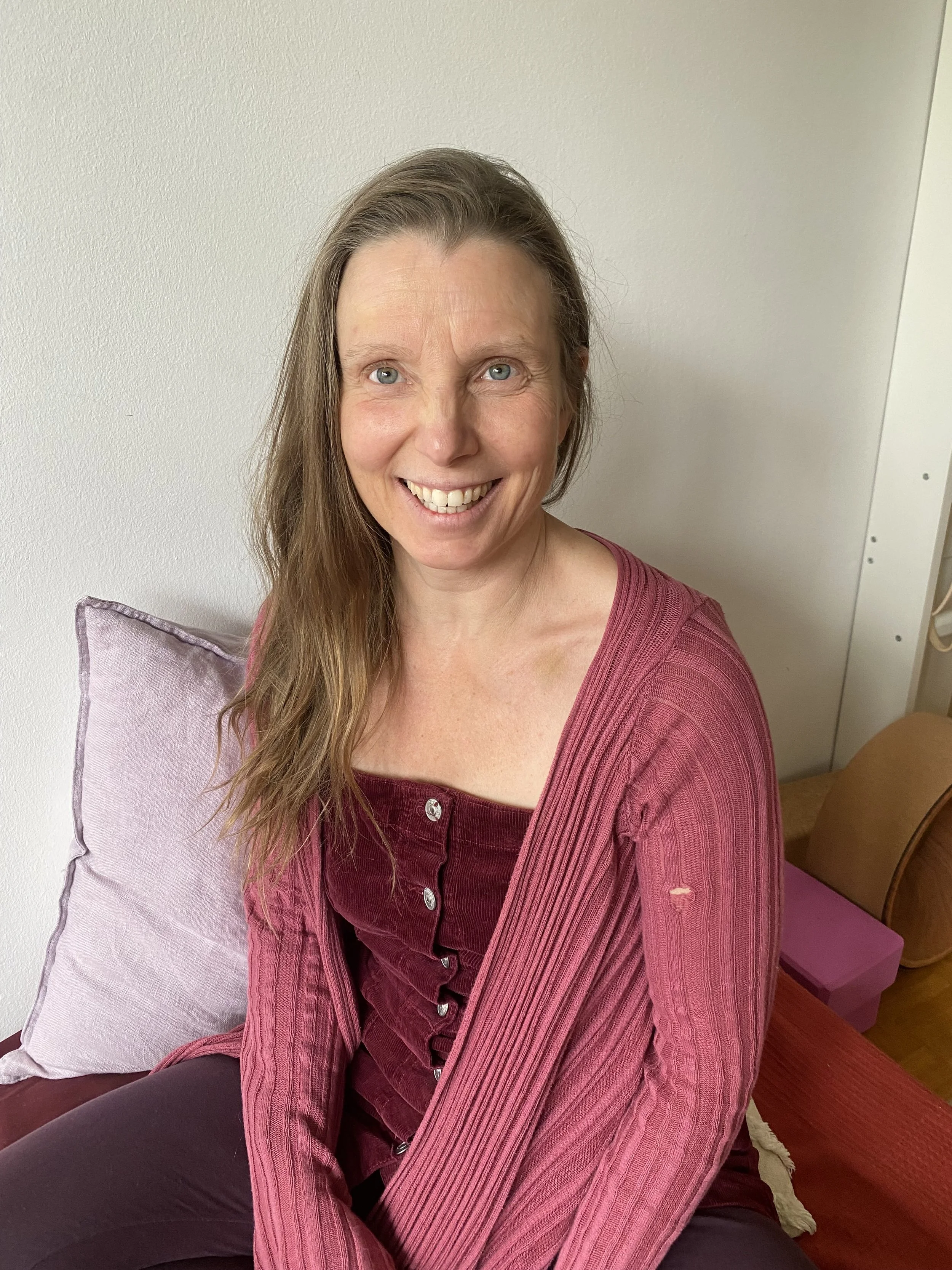 A woman with long brown hair, blue eyes, and a wide smile, sitting on a couch with pink and purple pillows, wearing a pink ribbed cardigan over a maroon top.