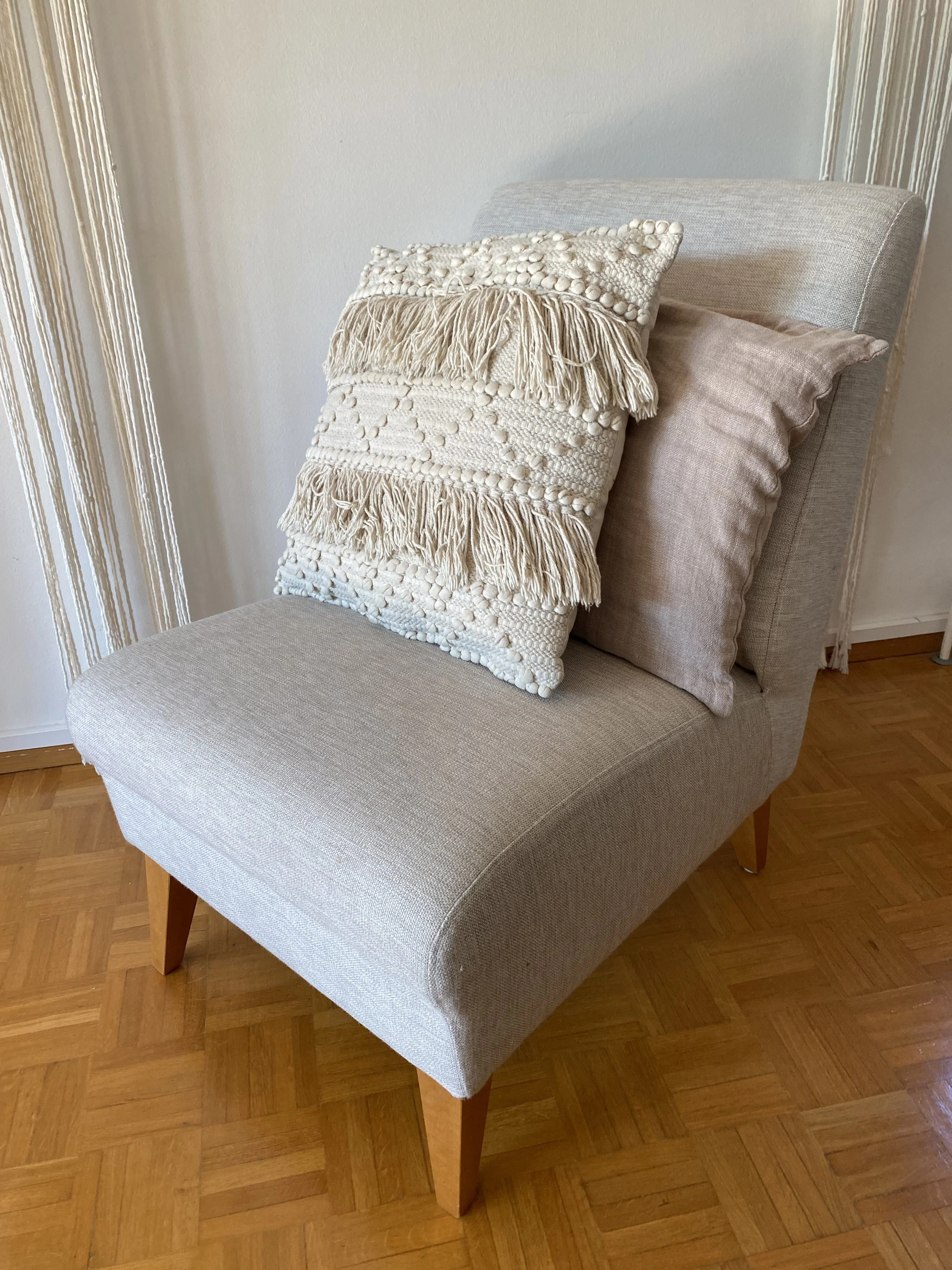 A beige upholstered chair with wooden legs, on a hardwood floor, with three decorative pillows placed on it.