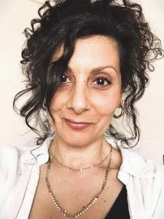 Close-up portrait of Amanda Murjan with dark curly hair, wearing a white top, a necklace, and earrings, against a plain background.