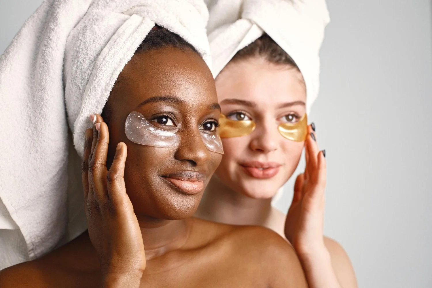 Two women with towels wrapped around their heads applying under-eye patches in a skincare routine.