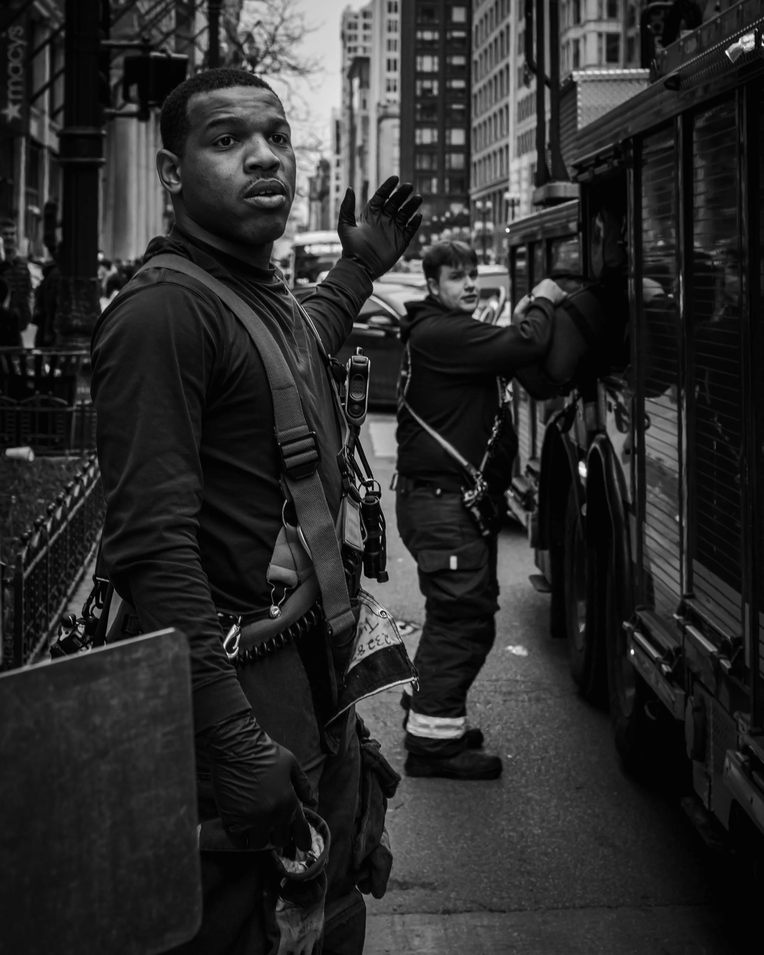Two police officers in tactical gear working on the street, one looking at the camera and the other opening a vending machine, with a city street and tall buildings in the background. THE COLOR OF FIRE. RJ CLARKE