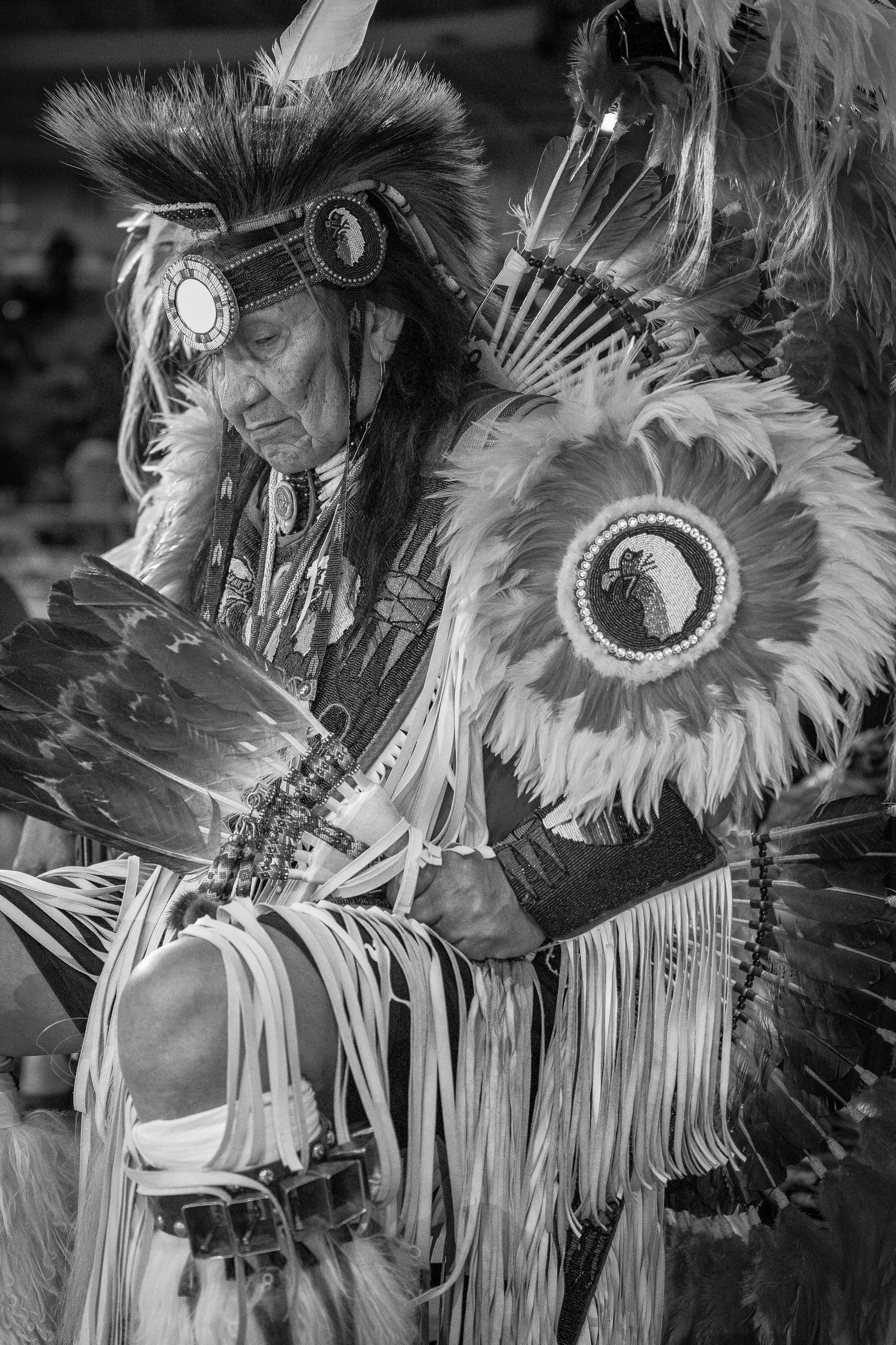 A Native American man dressed in traditional ceremonial attire with feathered headdress, face paint, and beadwork, engaged in a cultural dance or ritual. THE LAST DANCE. RJ CLARKE