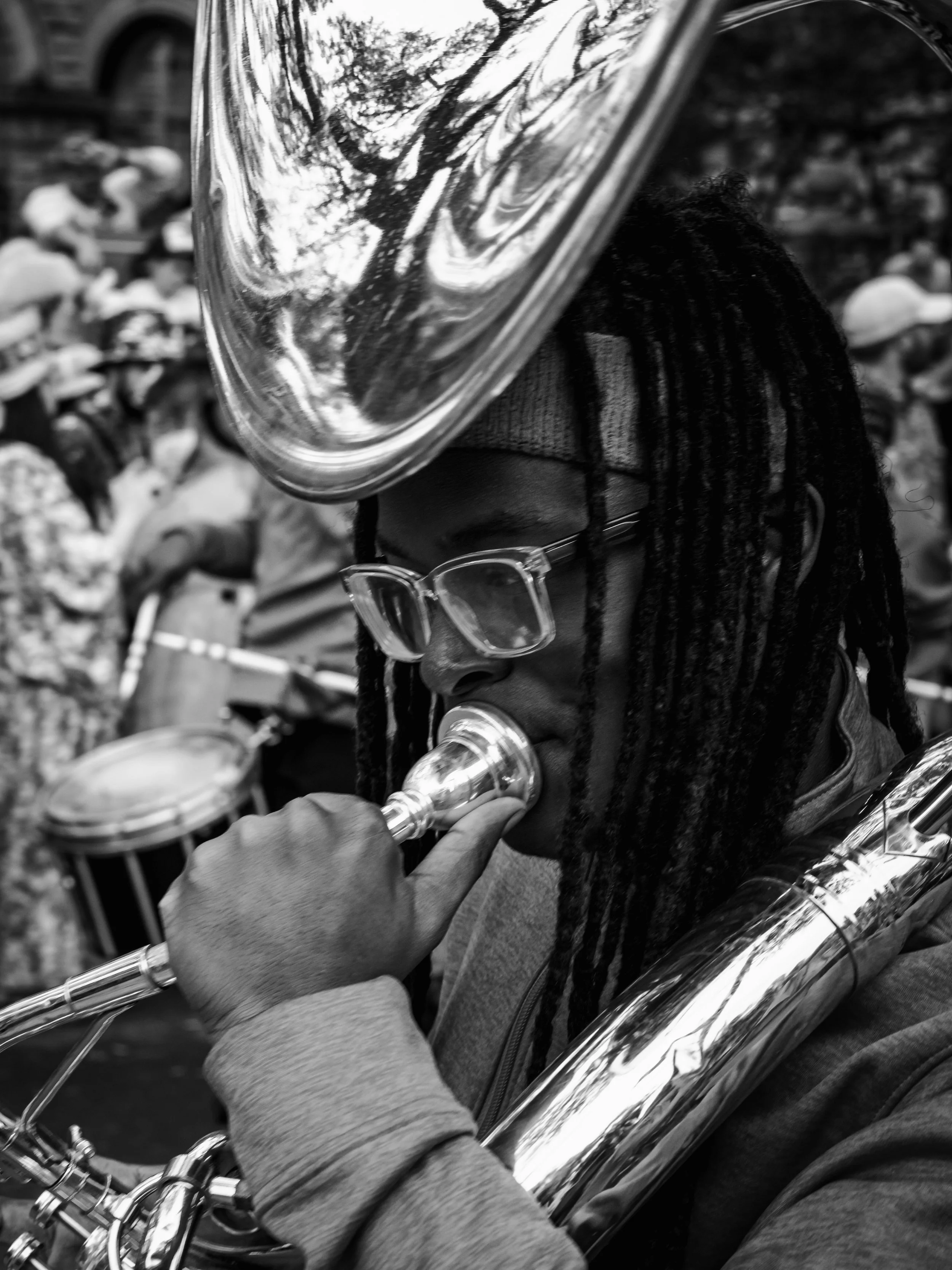 A person with dreadlocks wearing glasses and a sweater playing a trumpet.