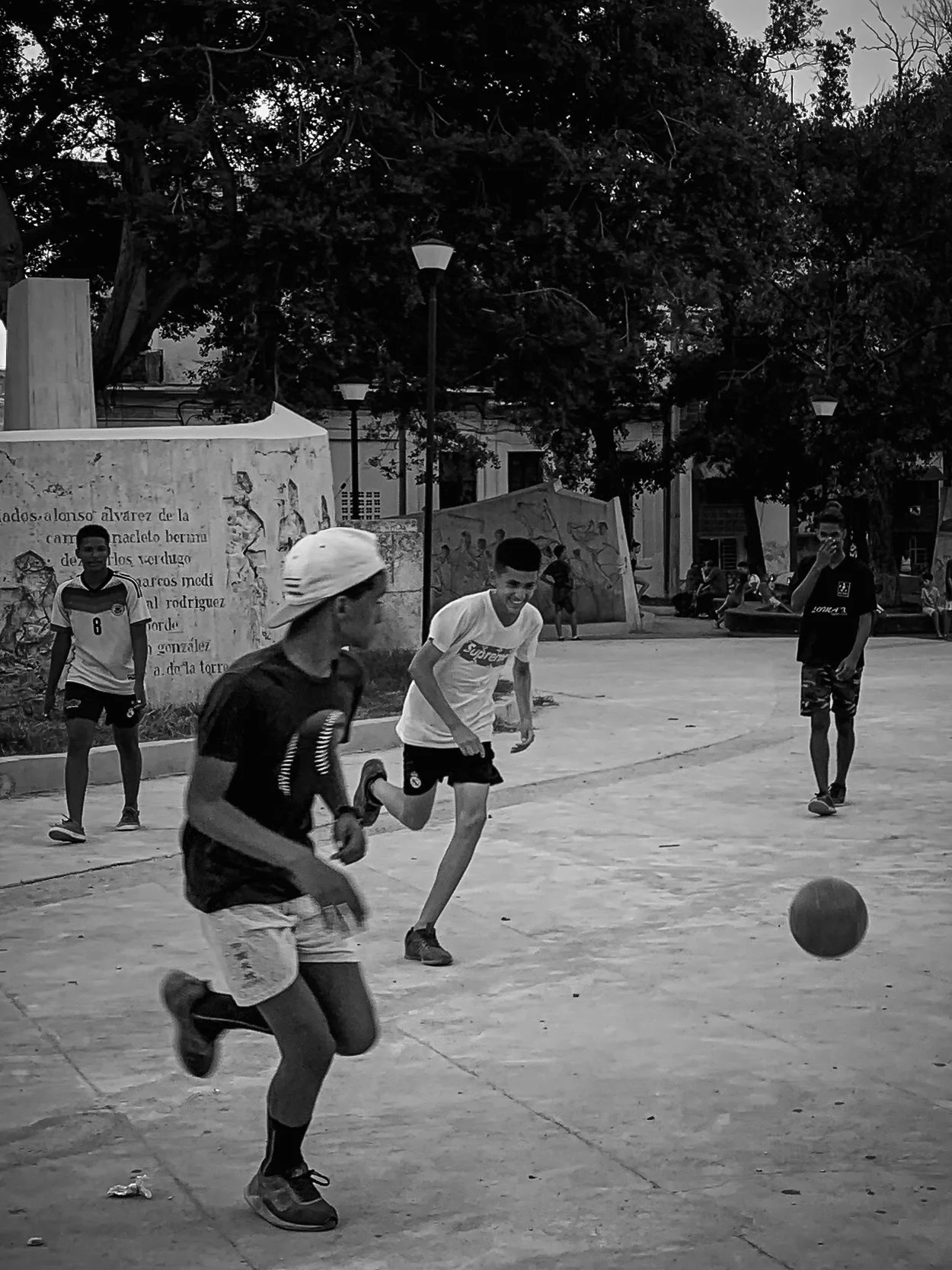 Children playing soccer in a park on a sunny day, with trees, benches, and a monument with text in the background.
