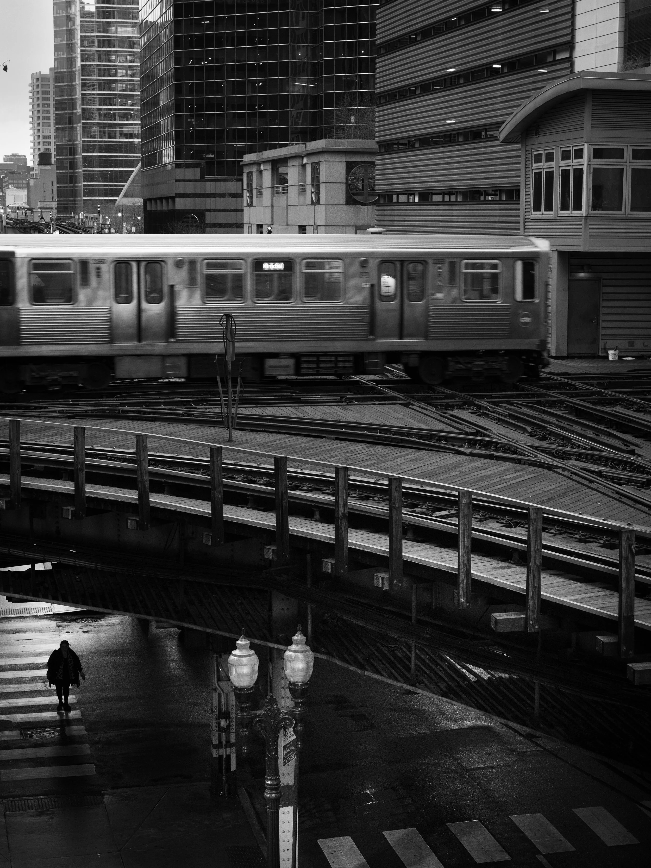 Black and white photo of an urban scene featuring a moving train on elevated tracks, with modern glass skyscrapers and buildings in the background. A pedestrian is crossing the street below, and a streetlamp with ornate design is visible in the foreg