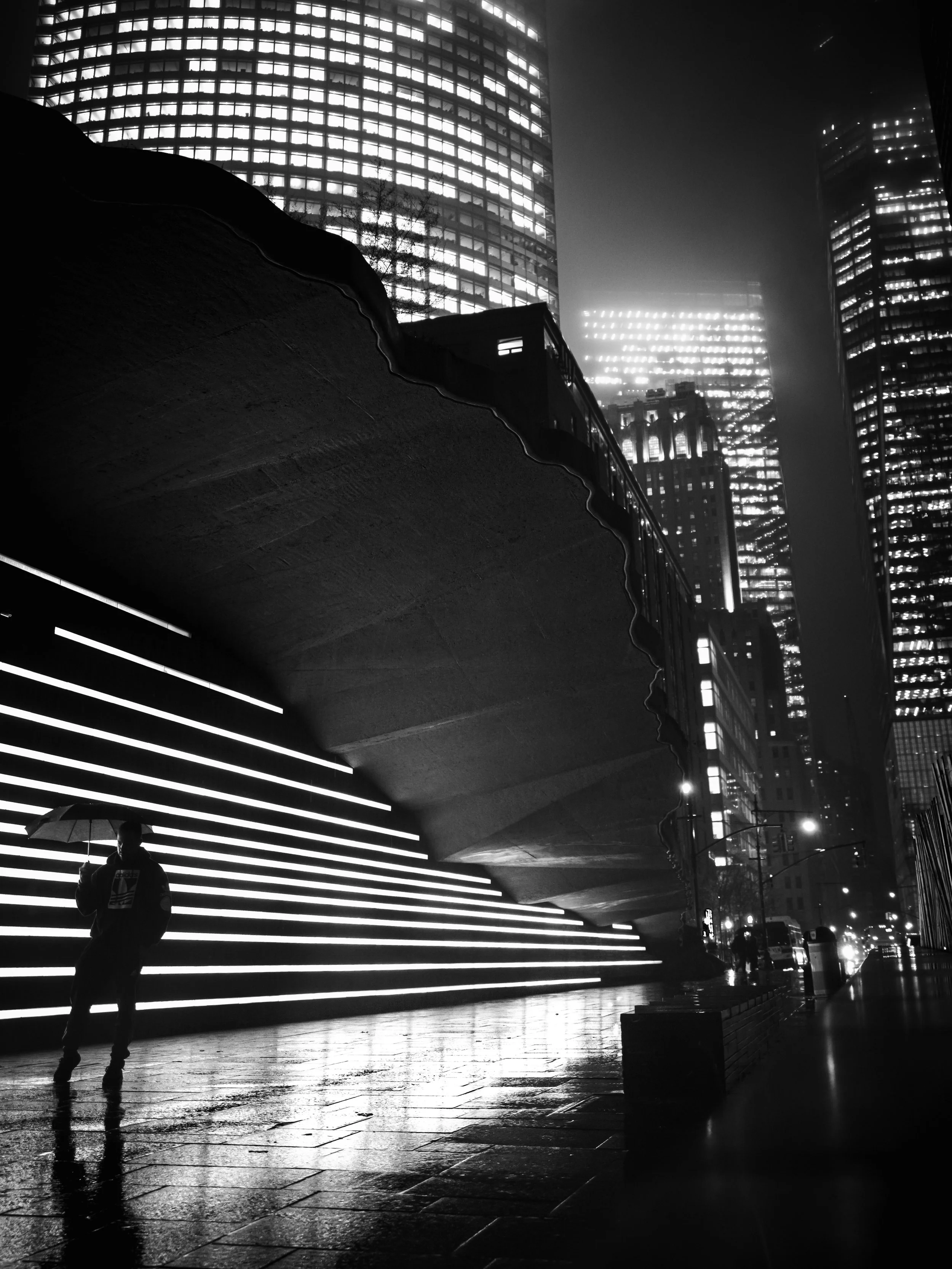 A black and white photograph of a cityscape at night, featuring illuminated skyscrapers, a person holding an umbrella walking on wet pavement, and a modern structure with horizontal light strips, possibly a bridge or walkway, in the foreground.