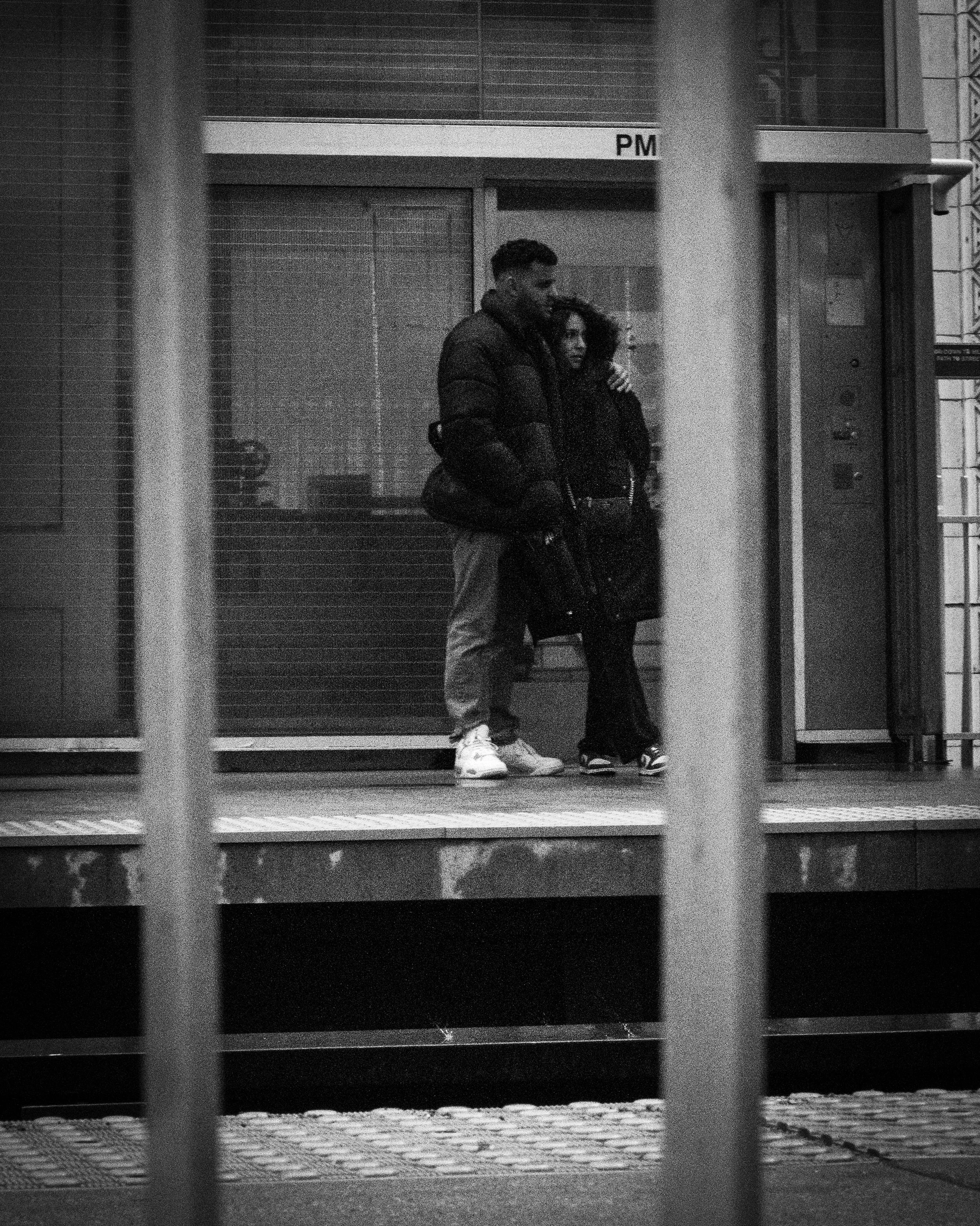 Black and white photo of a man and woman standing close together outside a building, with metal bars in the foreground.