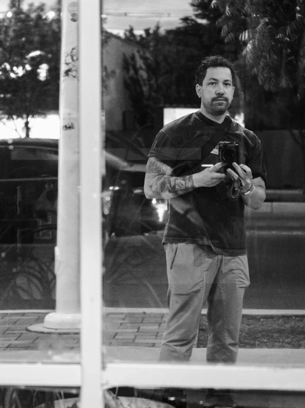 A man taking a selfie in a mirror, holding a camera, outdoors during evening or night, with trees and cars in the background, reflected through a window.  RJ CLARKE