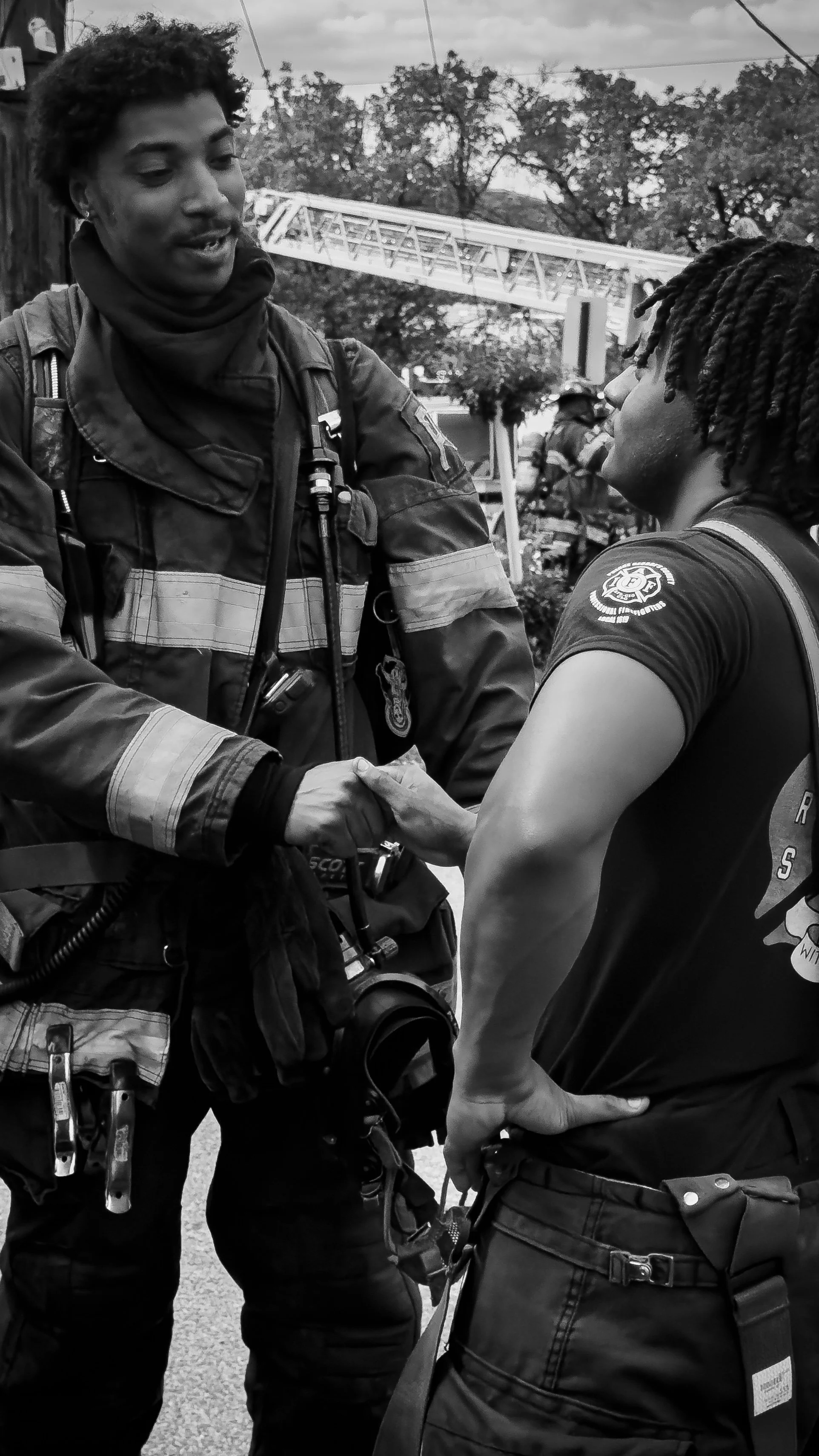 Two emergency responders shaking hands at an outdoor event, with trees and a fire truck ladder in the background. THE COLOR OF FIRE. RJ CLARKE. 