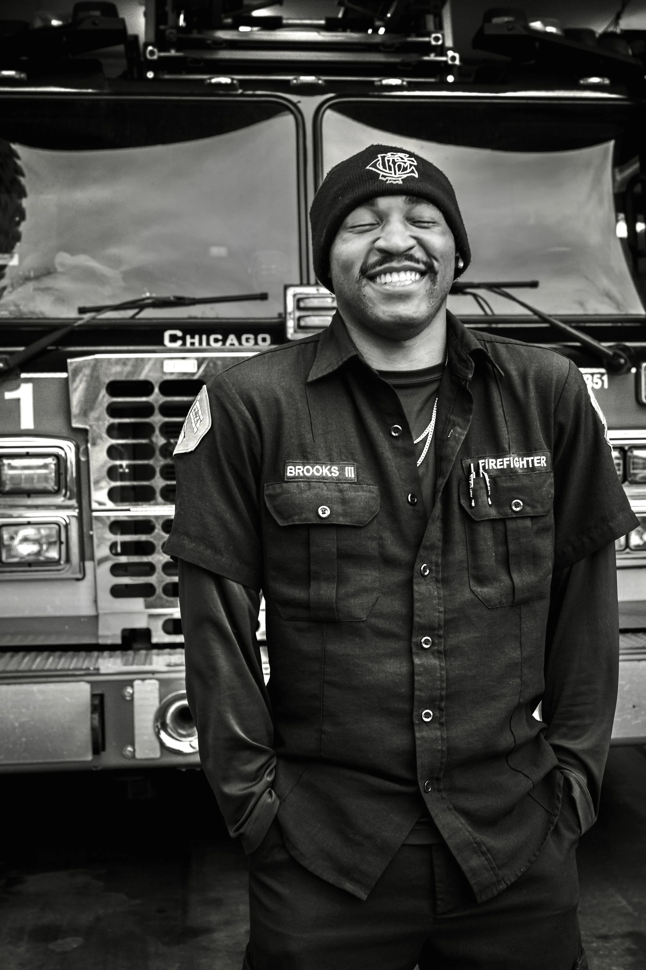 A smiling firefighter standing in front of a fire truck. THE COLOR OF FIRE. RJ Clarke