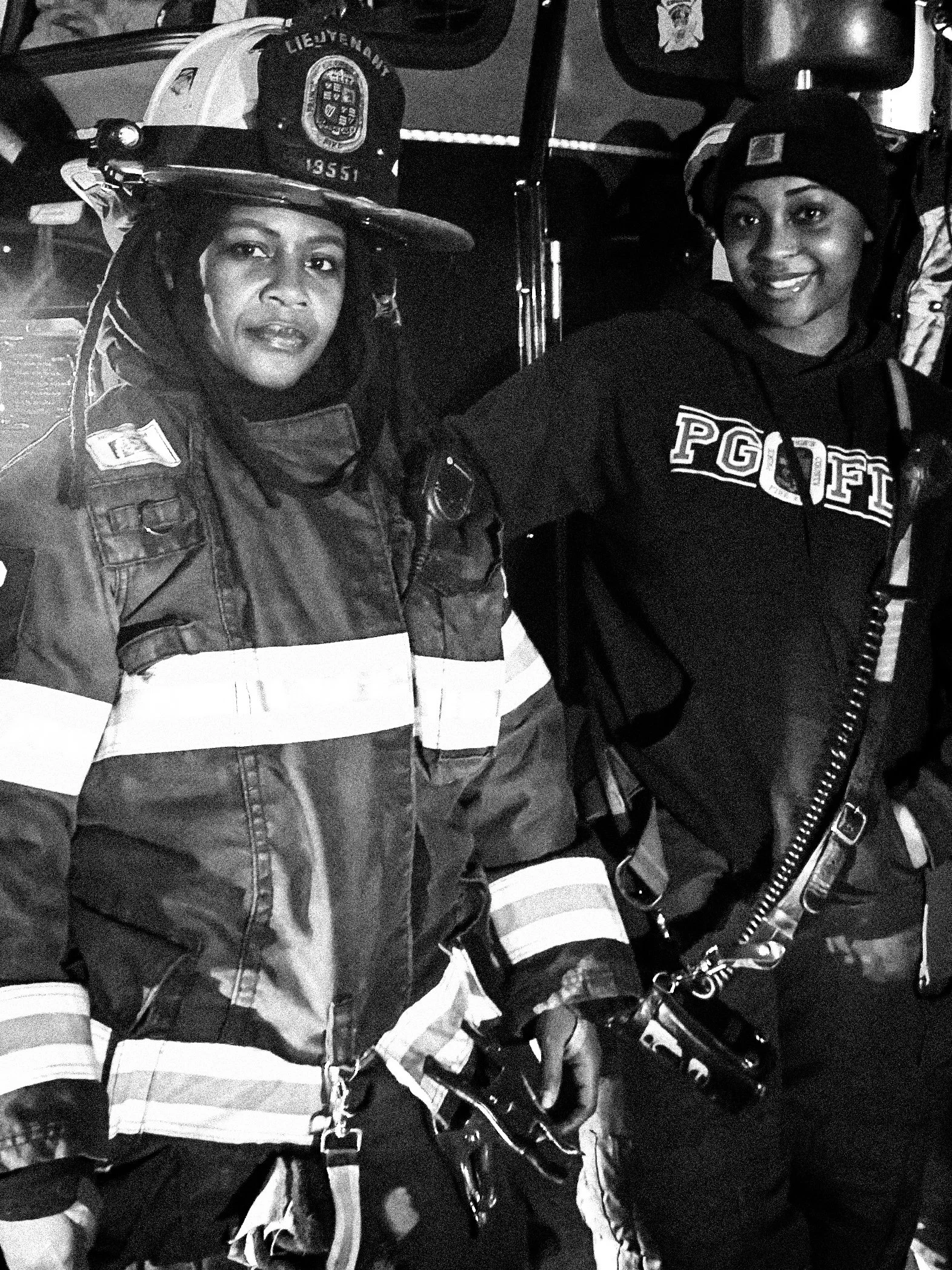 Two female firefighters in uniform, wearing helmets with patches, capturing a selfie at night using a radio device, with a fire truck in the background. THE COLOR OF FIRE. RJ CLARKE