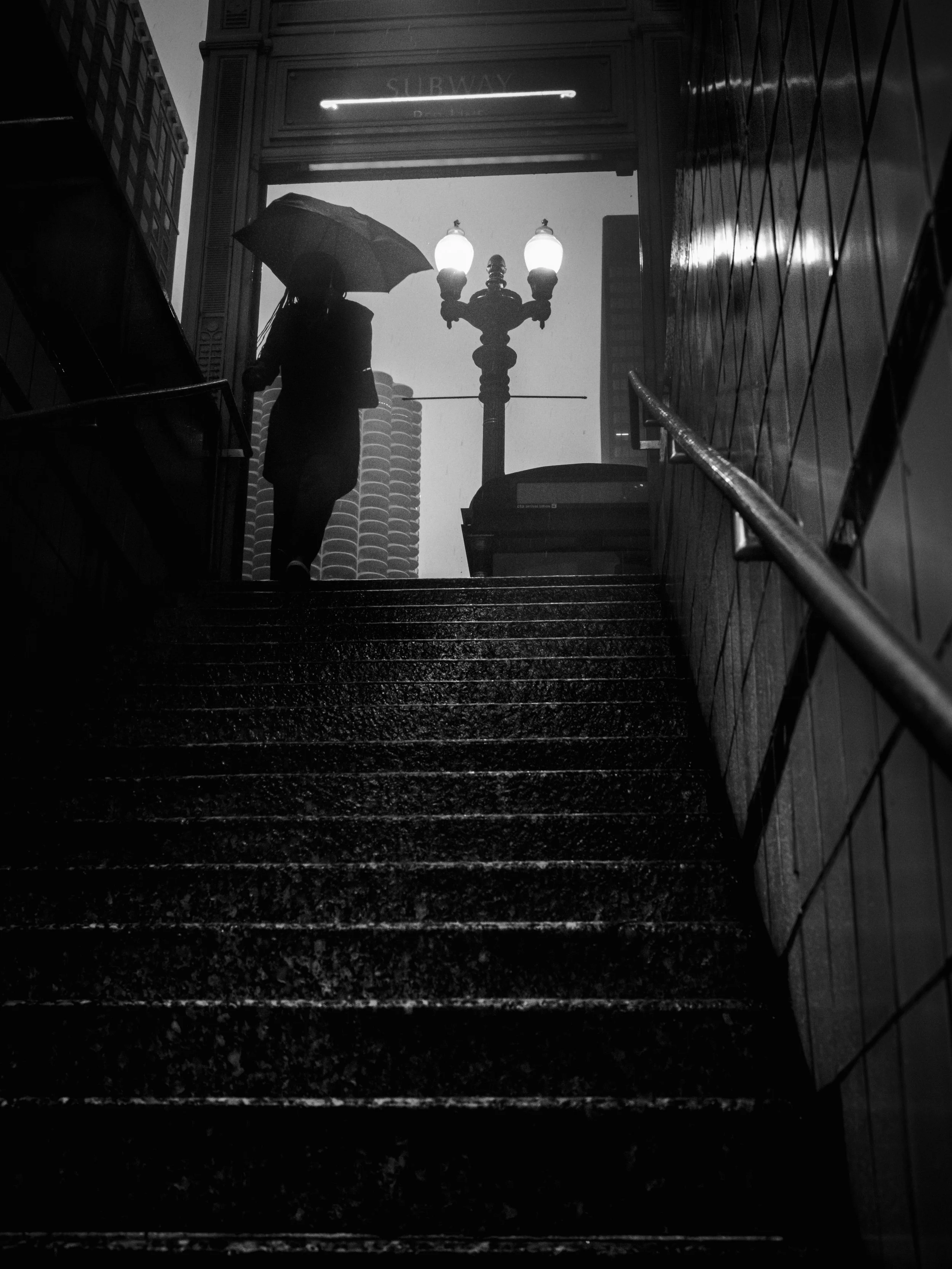 A silhouette of a woman holding an umbrella walking up stairs at night, with a lit street lamp in the background and tall buildings around.