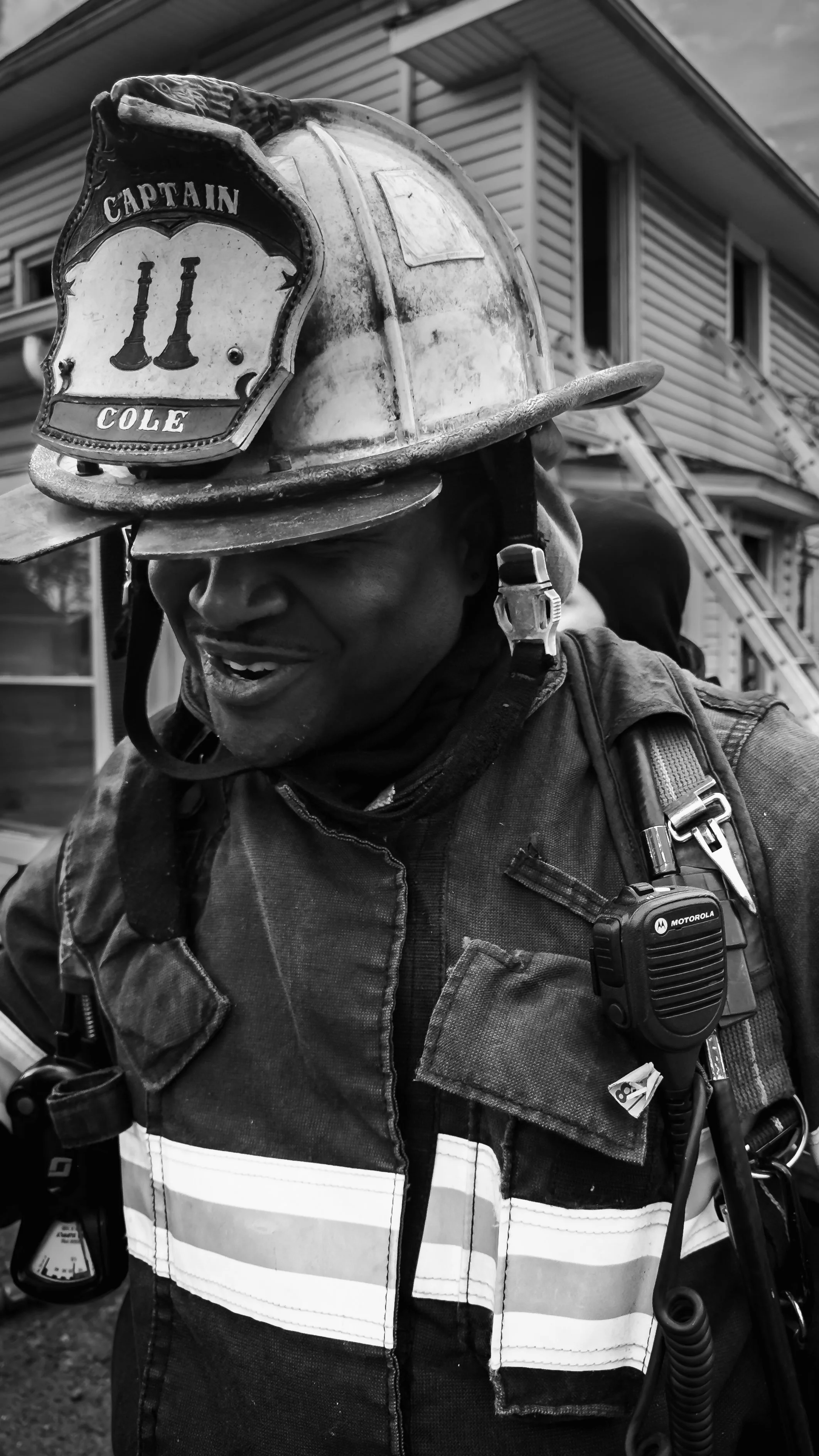 A firefighter wearing a helmet with a badge that says 'Captain Cole' and firefighting gear, smiling and talking to someone outdoors in front of a house. THE COLOR OF FIRE. RJ CLARKE
