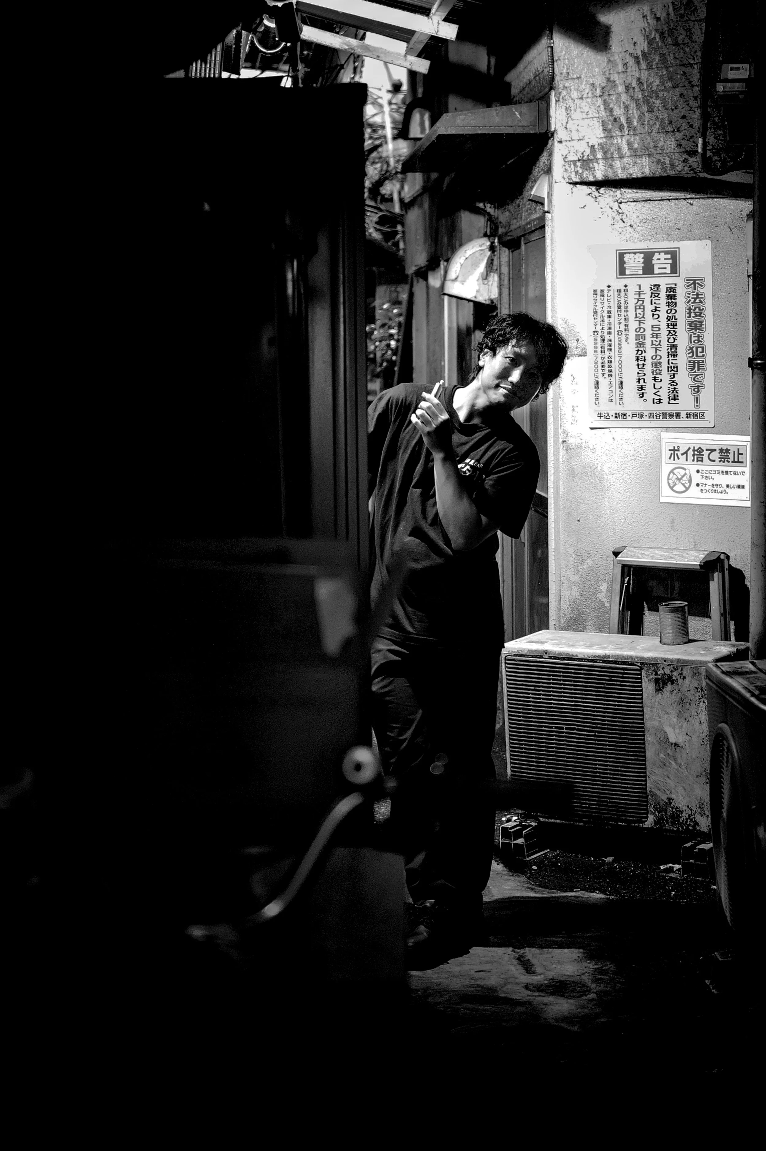 A young man standing outdoors at night, with a smile, holding a cigarette, next to a wall with Japanese warning signs, surrounded by urban elements.