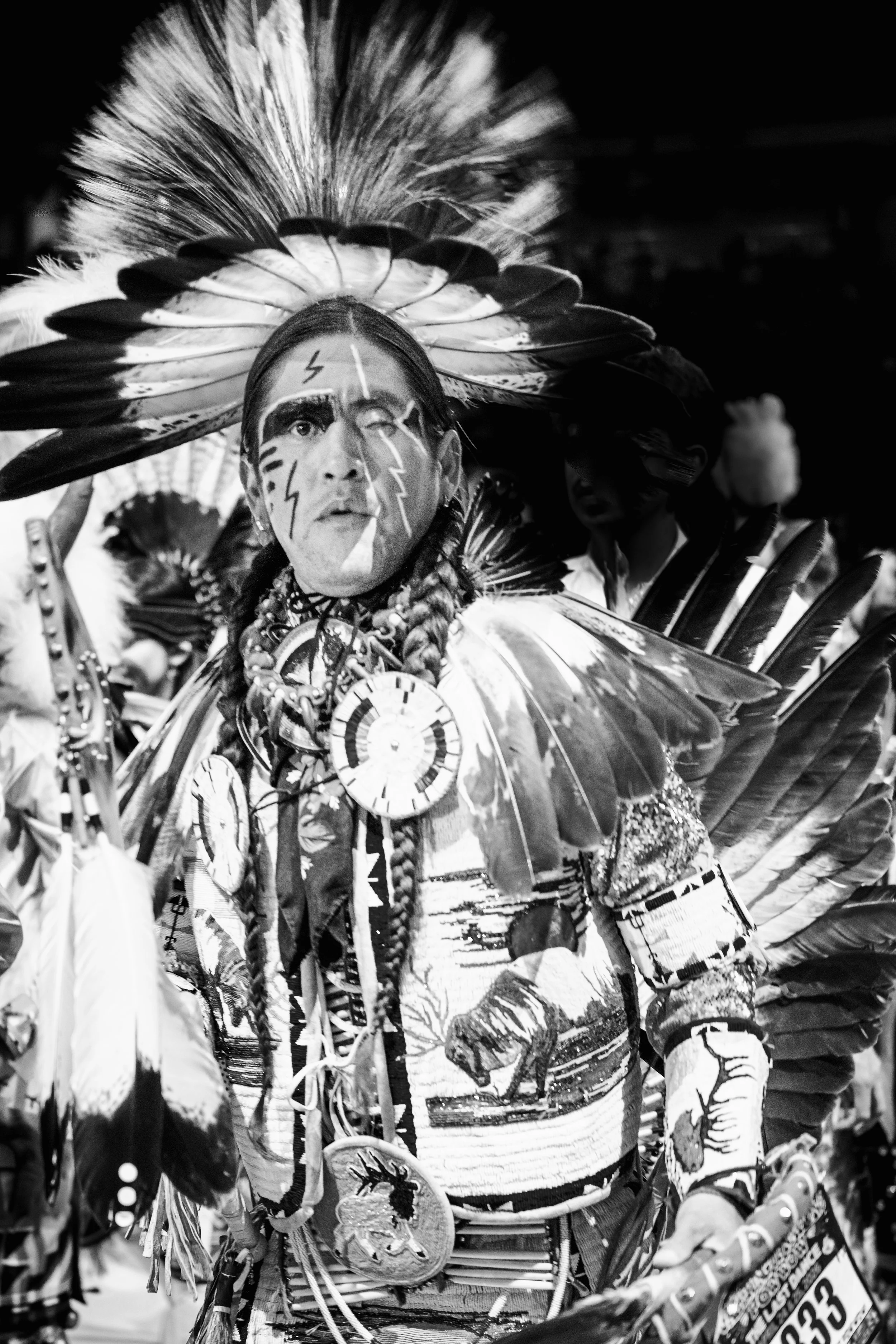 A person dressed in traditional indigenous attire, with face paint and a large headdress made of feathers, participating in a cultural event or parade. THE LAST DANCE. RJ CLARKE