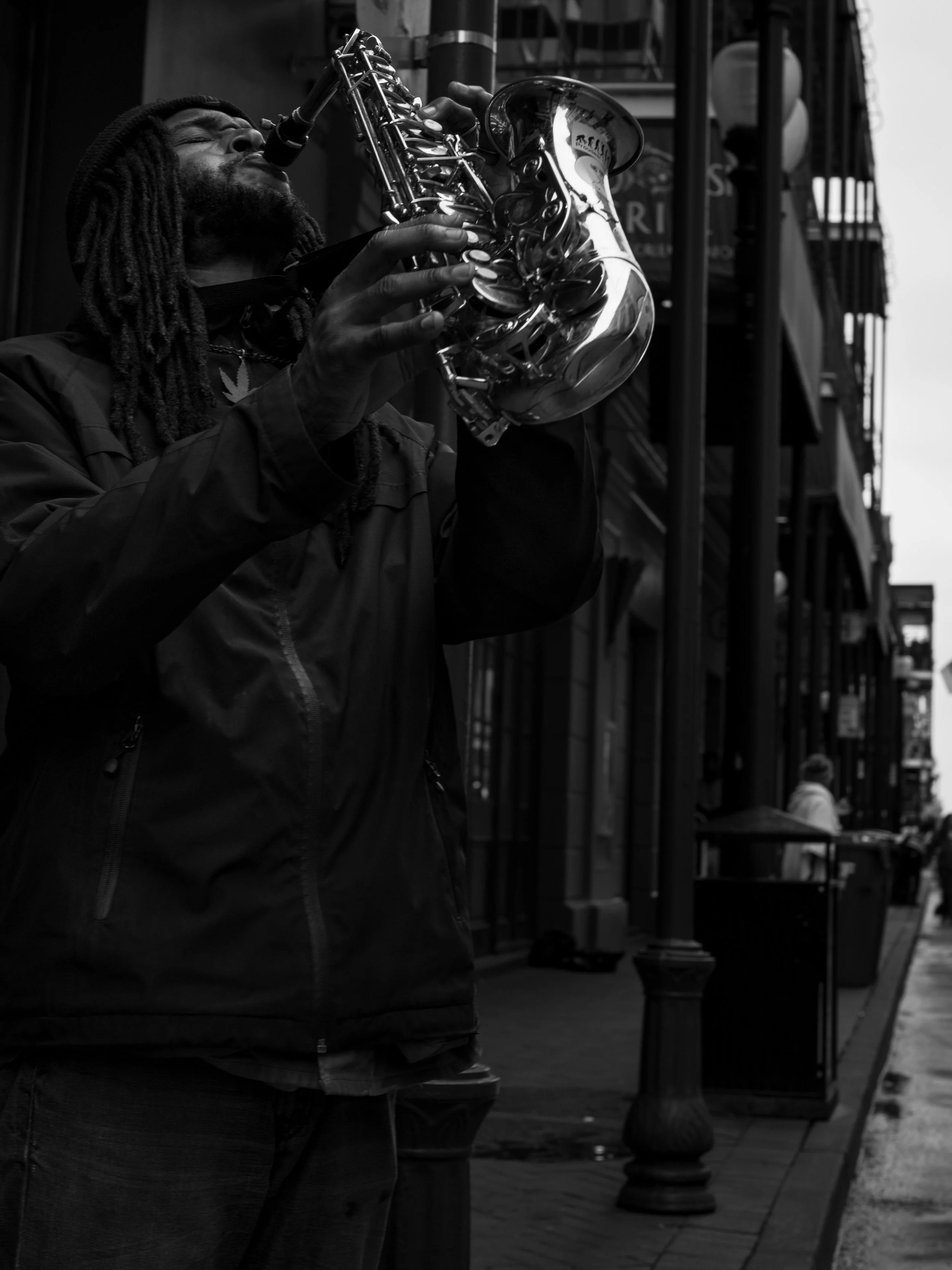 A man with dreadlocks playing a saxophone on a city street, black and white photograph.