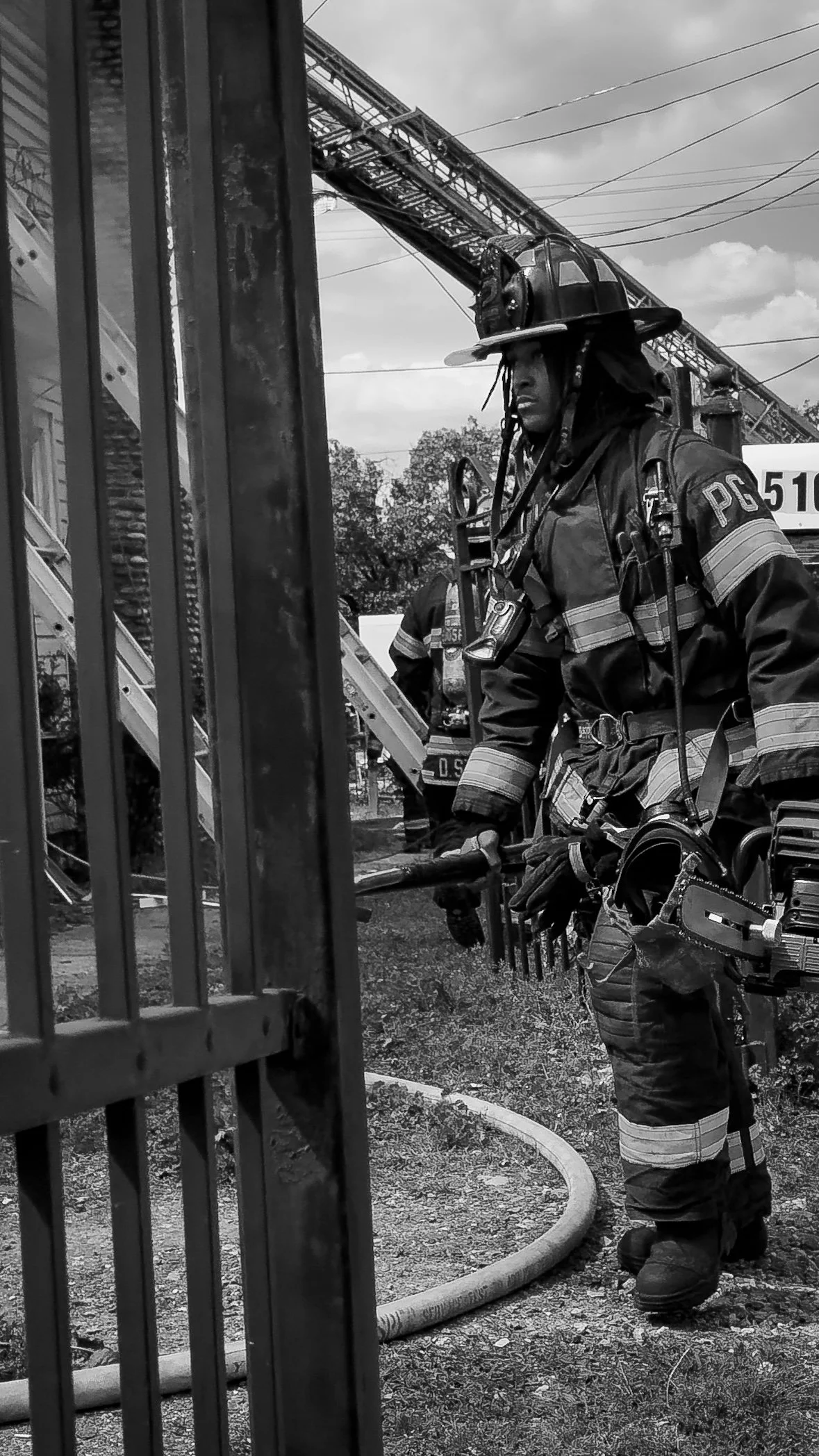 Black and white photo of a firefighter in full gear, including a helmet and protective suit, standing near a metal gate at a fire scene with a fire truck and equipment in the background. THE COLOR OF FIRE. RJ CLARKE
