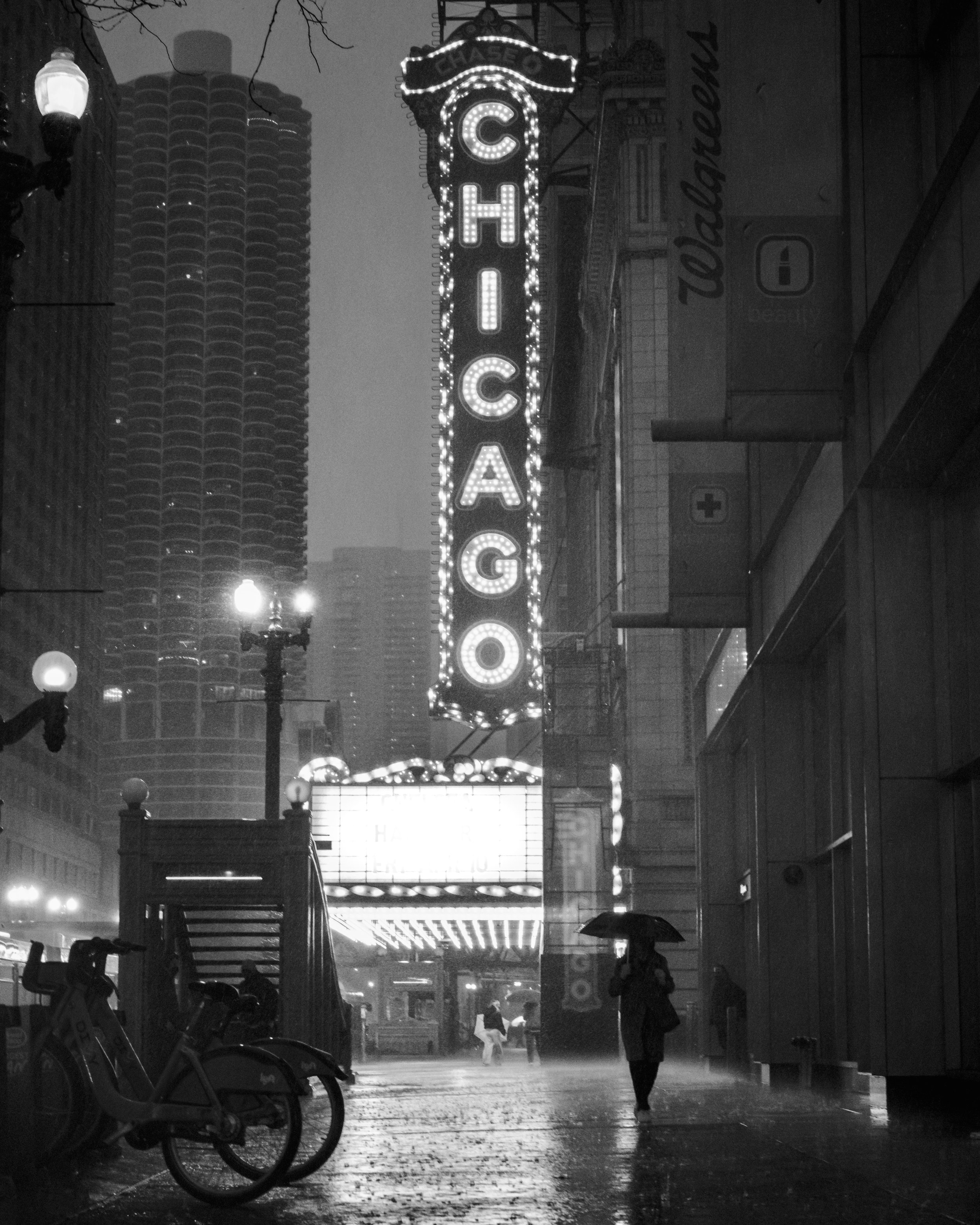 A rainy city street scene at night with a large, illuminated casino sign reading 'CHICAGO' hanging above the sidewalk. People are walking with umbrellas, and bicycles are parked on the side. Street lamps illuminate the wet pavement, and tall building
