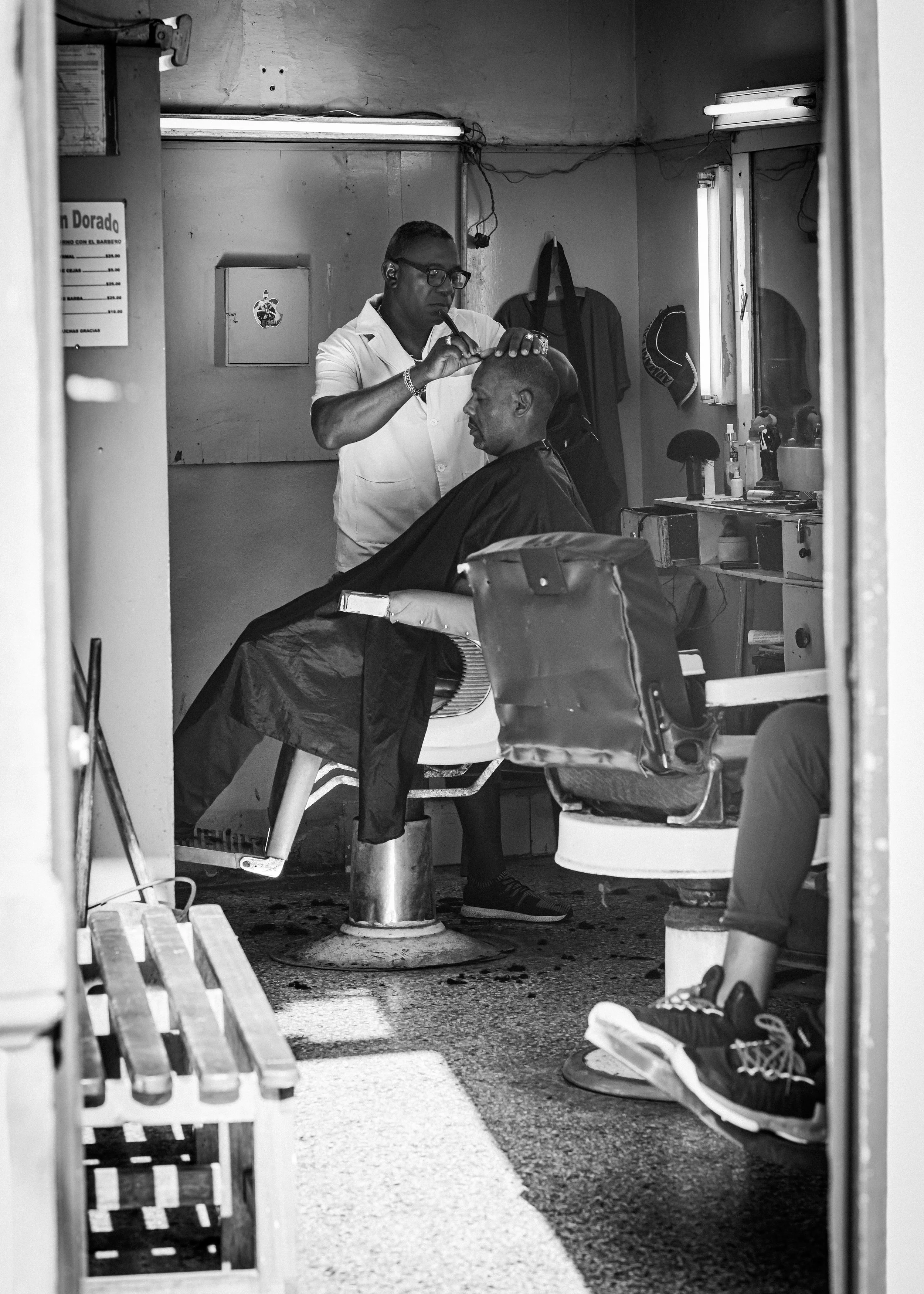 A barbershop scene in black and white showing a barber trimming a customer's hair. The customer is seated with a cape, while the barber is focused, wearing glasses and a white shirt. The setting includes a mirror, a window with blinds, and various gr