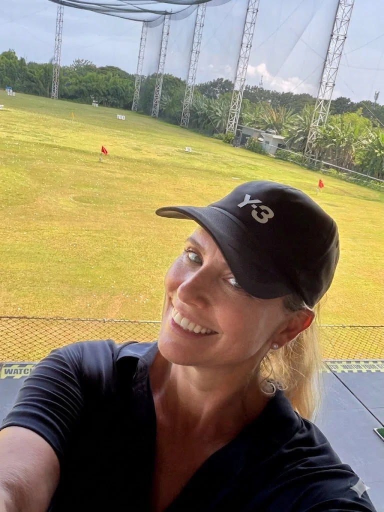 Frau im schwarzen Cap mit weißem Logo, die auf einer Driving Range steht, umgeben von Golfbahnen, Abschlagsmarkierungen und einem Netz, mit Bäumen im Hintergrund.