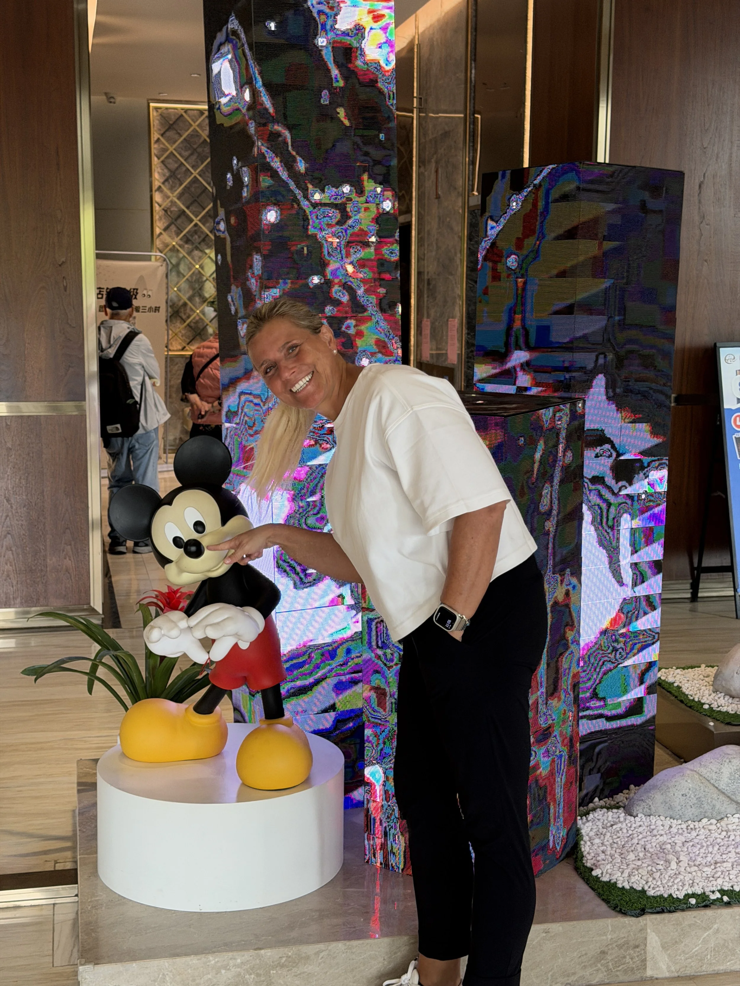 Eine lachende Frau in weißem T-Shirt steht neben einer Mickey-Mouse-Figur auf einer runden weißen Ablage. Im Hintergrund sind bunte digitale Bildschirme und eine Wanddekoration mit Steinen und Pflanzen.