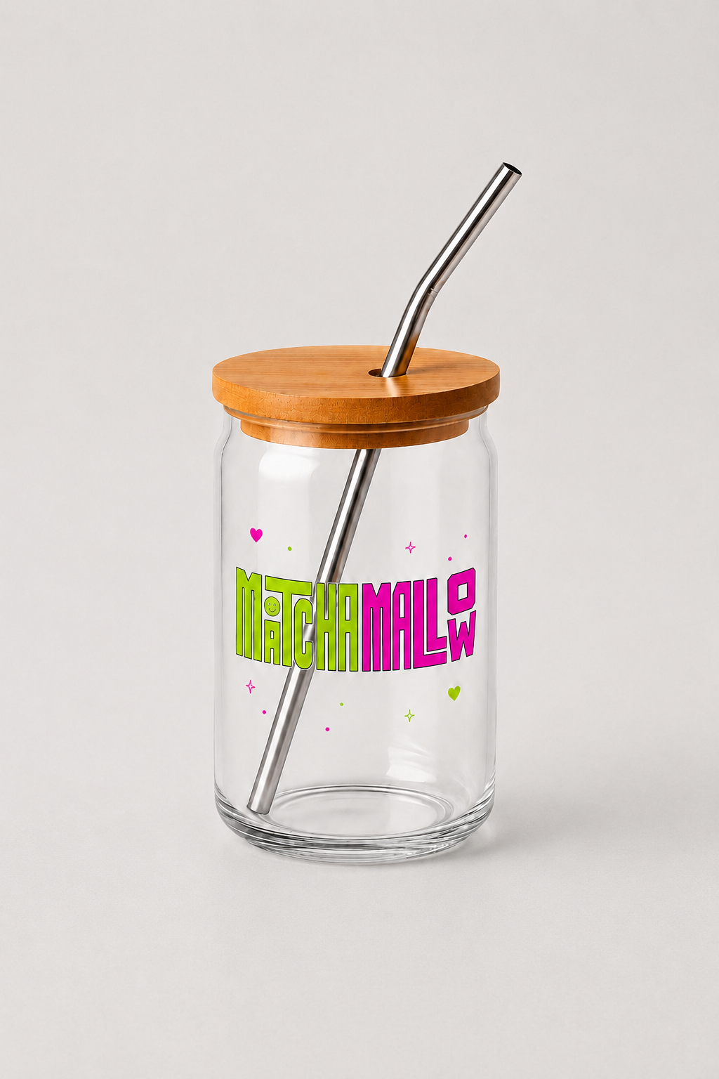 Ein transparentes Trinkglas mit einem Holzdeckel und einem Metallstrohhalm. Das Glas hat bunte Schriftzug "MatchaMallow" mit kleinen Herzen und Sternen.