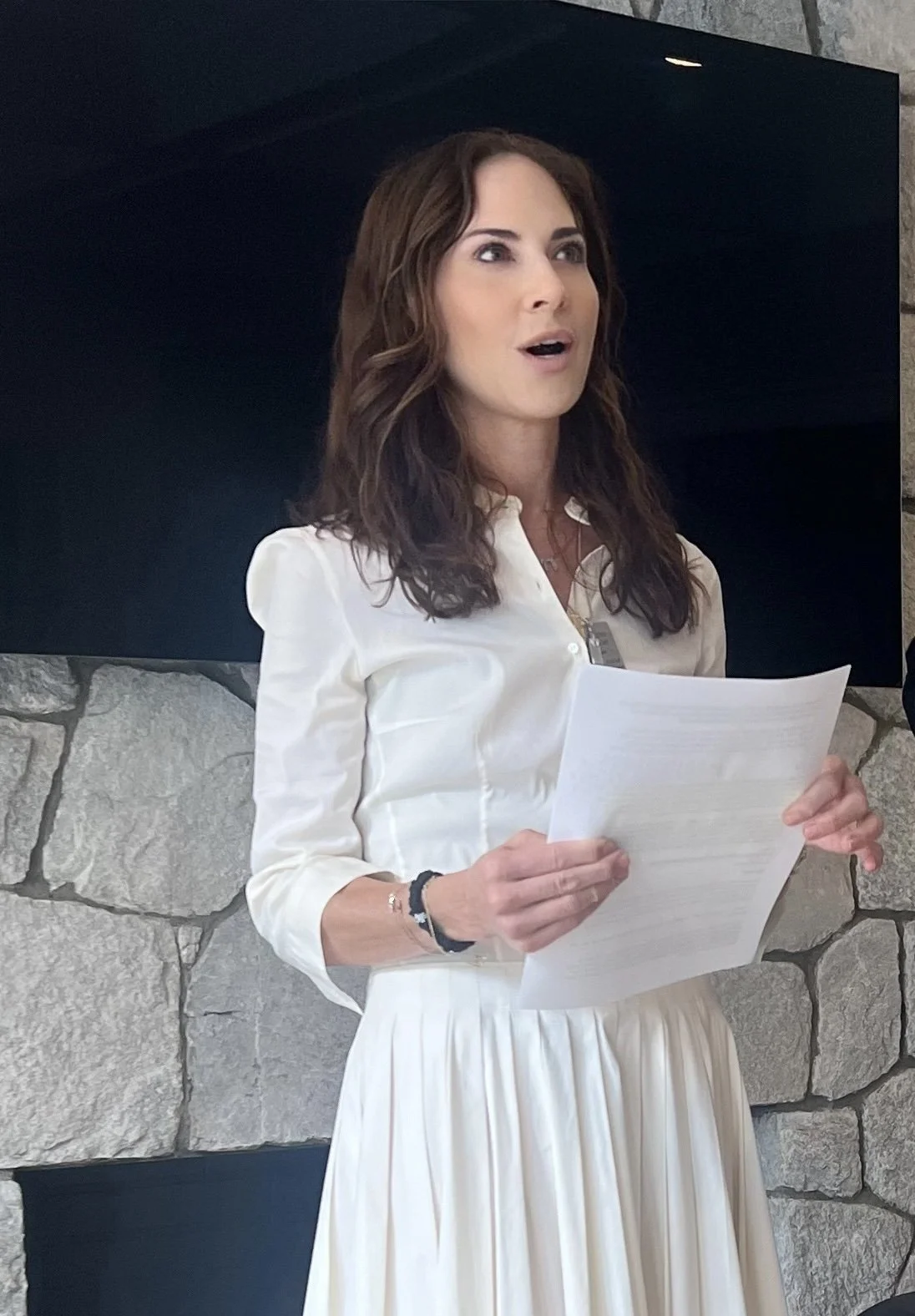 A woman with brown hair, wearing a white dress and holding papers, appears to be speaking or giving a presentation in front of a stone wall and a black background.