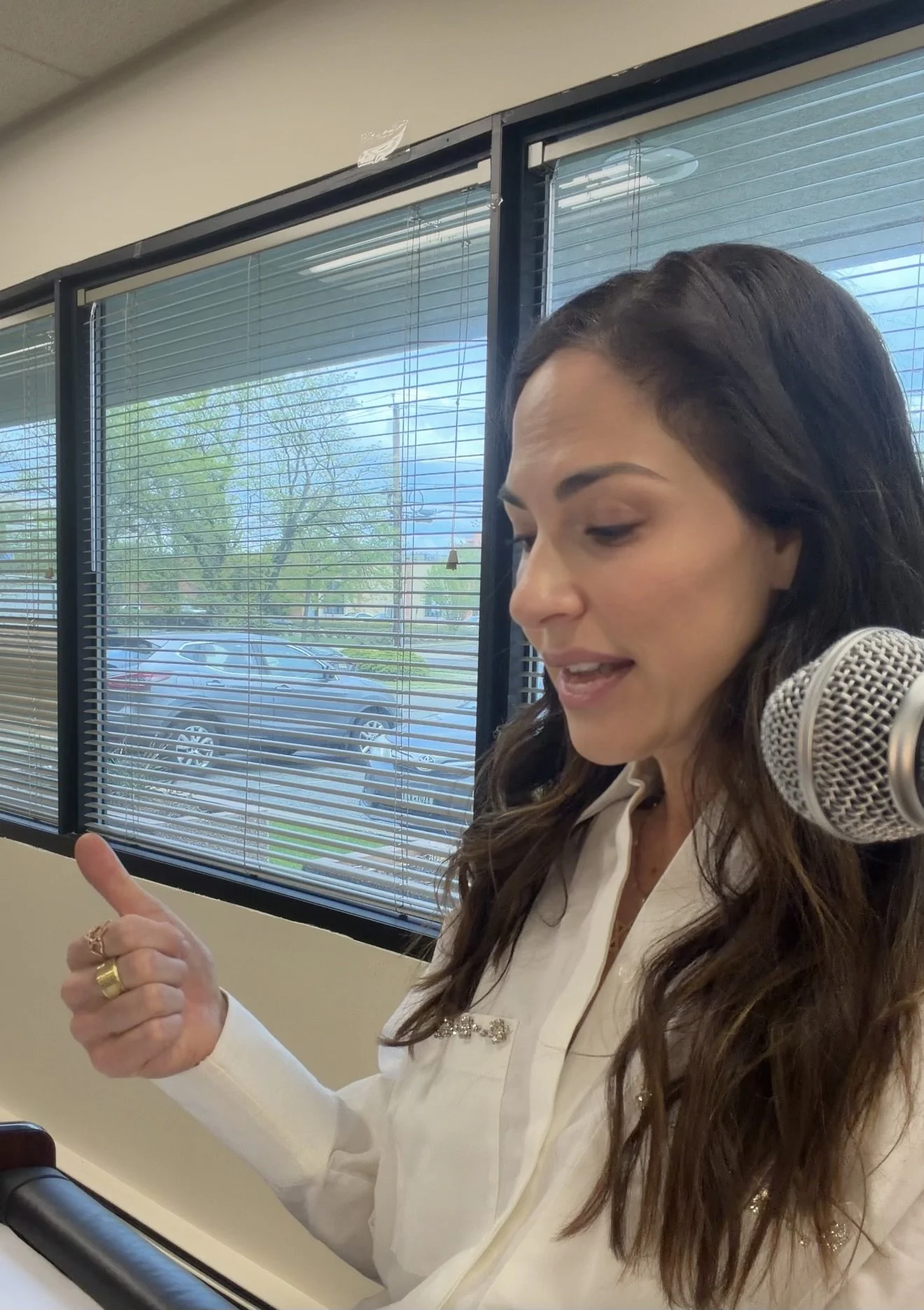 A woman with long dark hair, wearing a white blouse, is speaking into a microphone. She is gesturing with her left hand and has multiple rings on her fingers. Behind her are windows with blinds and a parking lot outside.