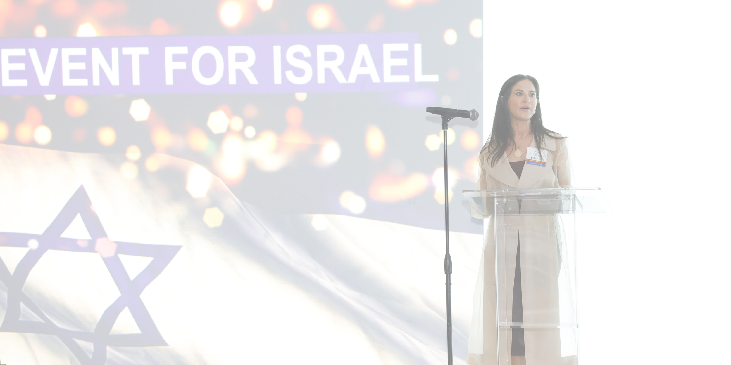 A woman with long dark hair, wearing a beige trench coat, stands at a clear podium with a microphone, speaking at an event. Behind her, a large screen displays the text 'EVENT FOR ISRAEL' with a graphic background of bokeh light effects and a geometric logo.
