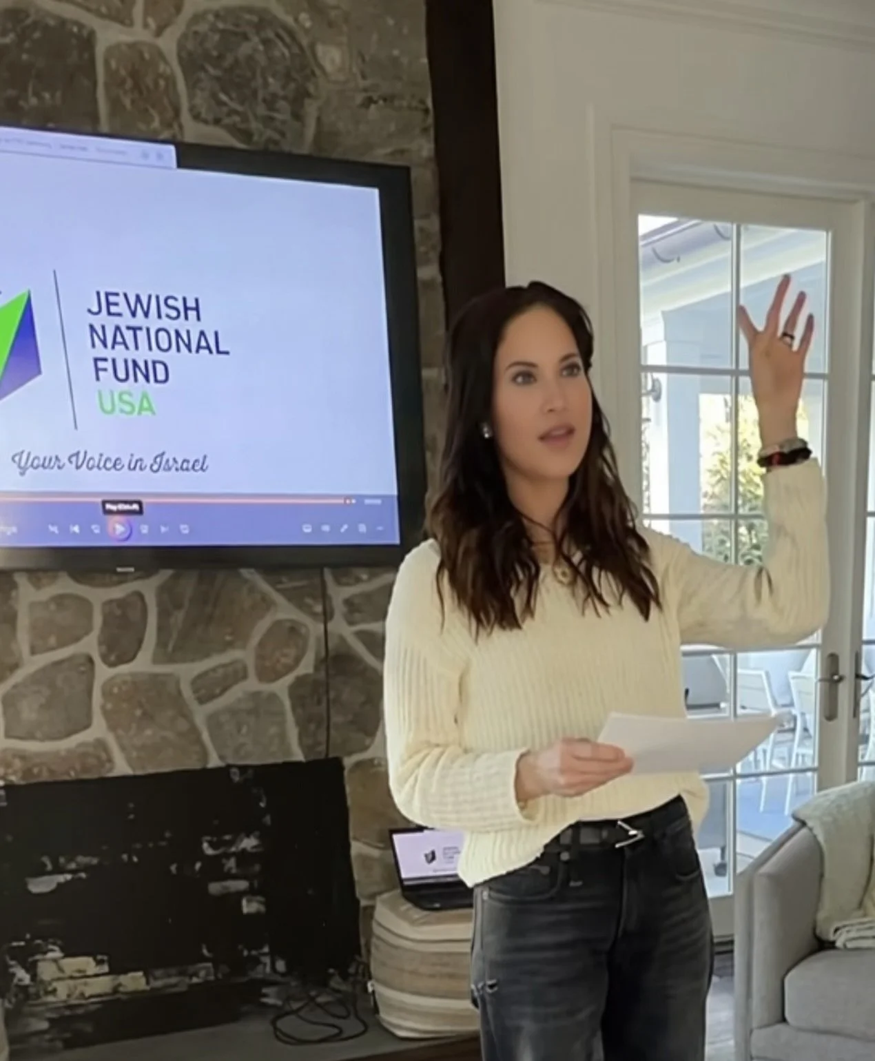 A woman with long dark hair and light skin, wearing a white sweater and black jeans, stands in front of a stone brick wall and a TV screen, holding a piece of paper in one hand and gesturing with her other hand during a presentation or speech.
