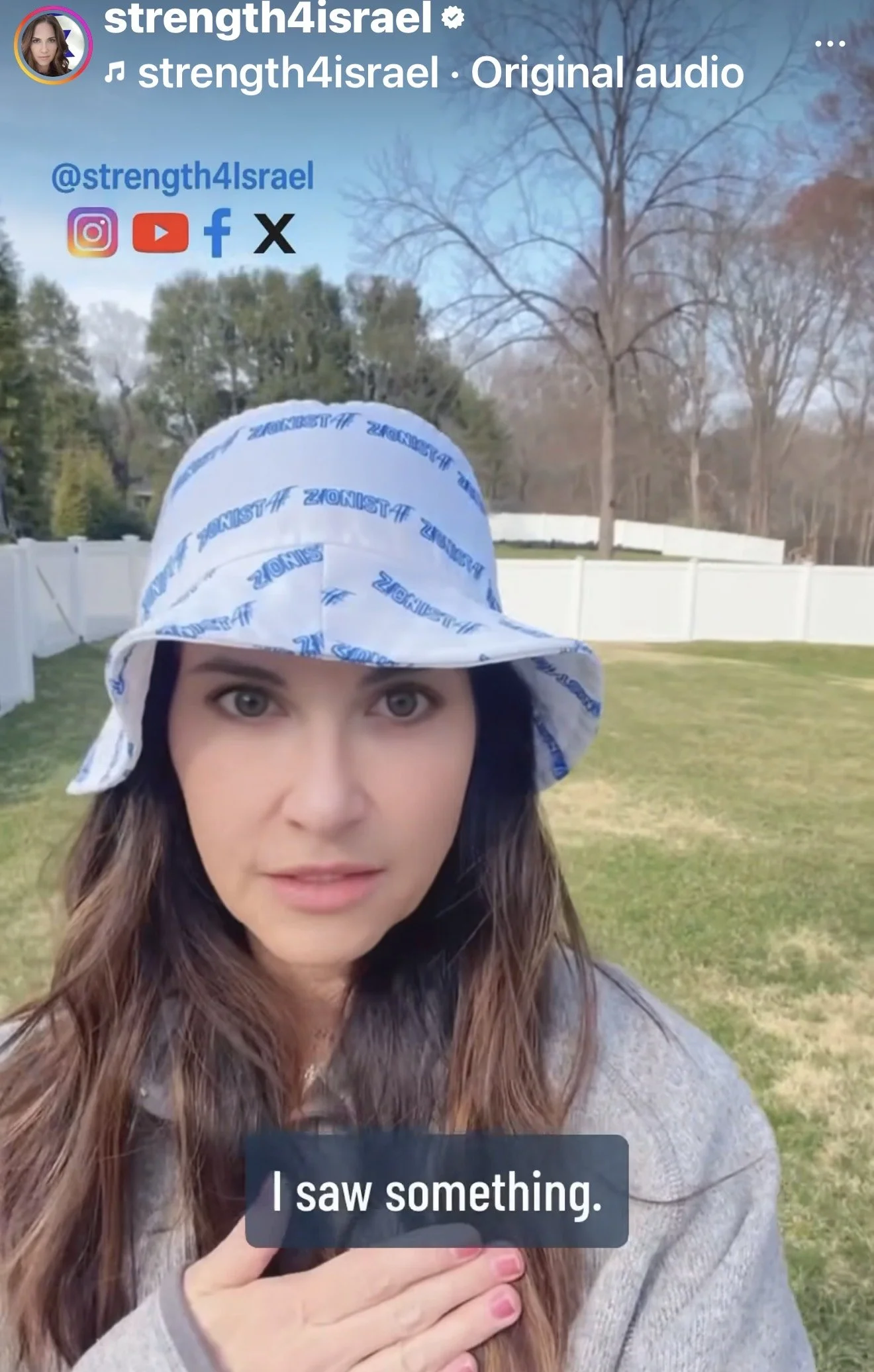 A woman with long brown hair wearing a white bandana with blue text and a gray hoodie, standing outdoors in a backyard with a white fence and trees in the background, holding a smartphone and looking into the camera. The image is a selfie with on-screen text that says "I saw something."