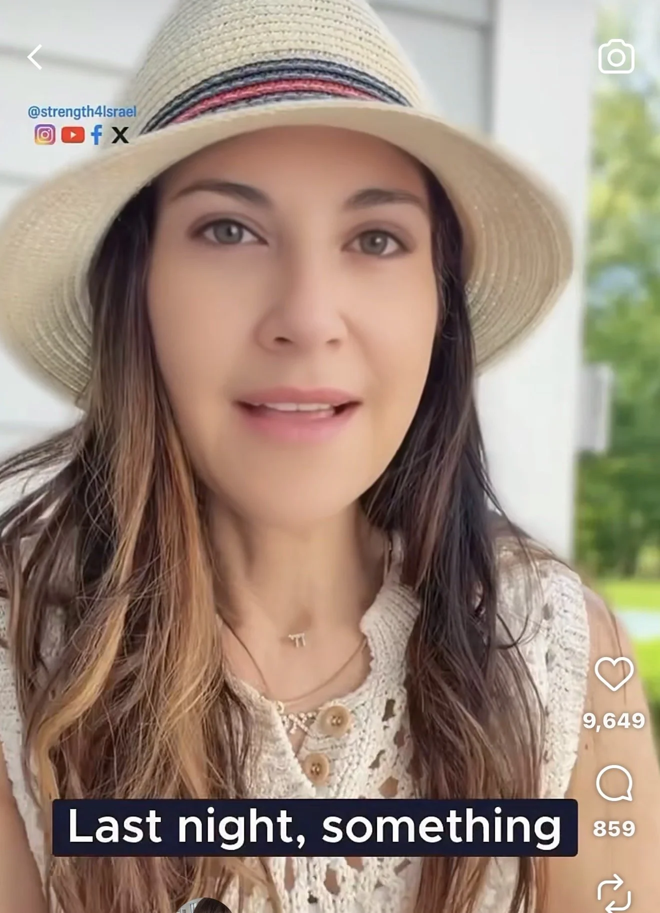 Close-up of a woman wearing a wide-brimmed straw hat with a colorful band, a lace top with buttons, and layered necklaces. She has long wavy hair and light-colored eyes, standing outdoors with greenery in the background.