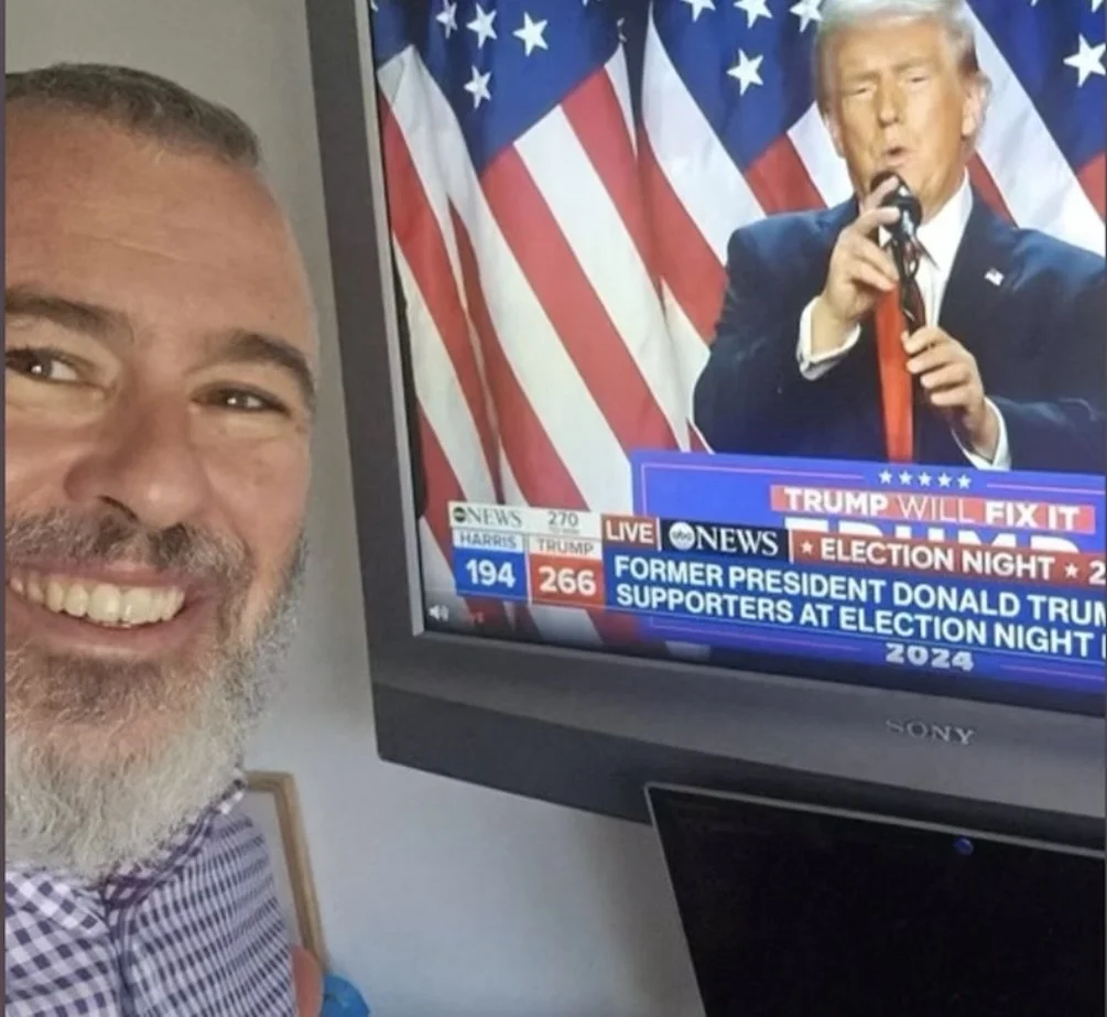 A man with a beard and short hair smiling at a camera, watching a TV screen showing former President Donald Trump speaking with American flags in the background.