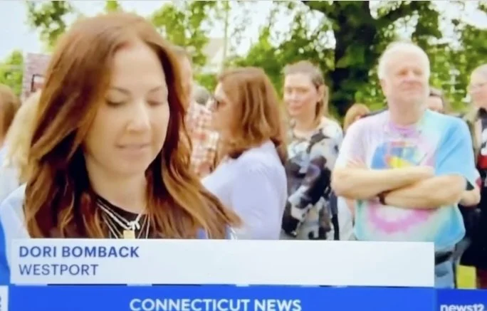 A woman with long red hair speaking on a news segment outdoors, with a crowd of people in the background.