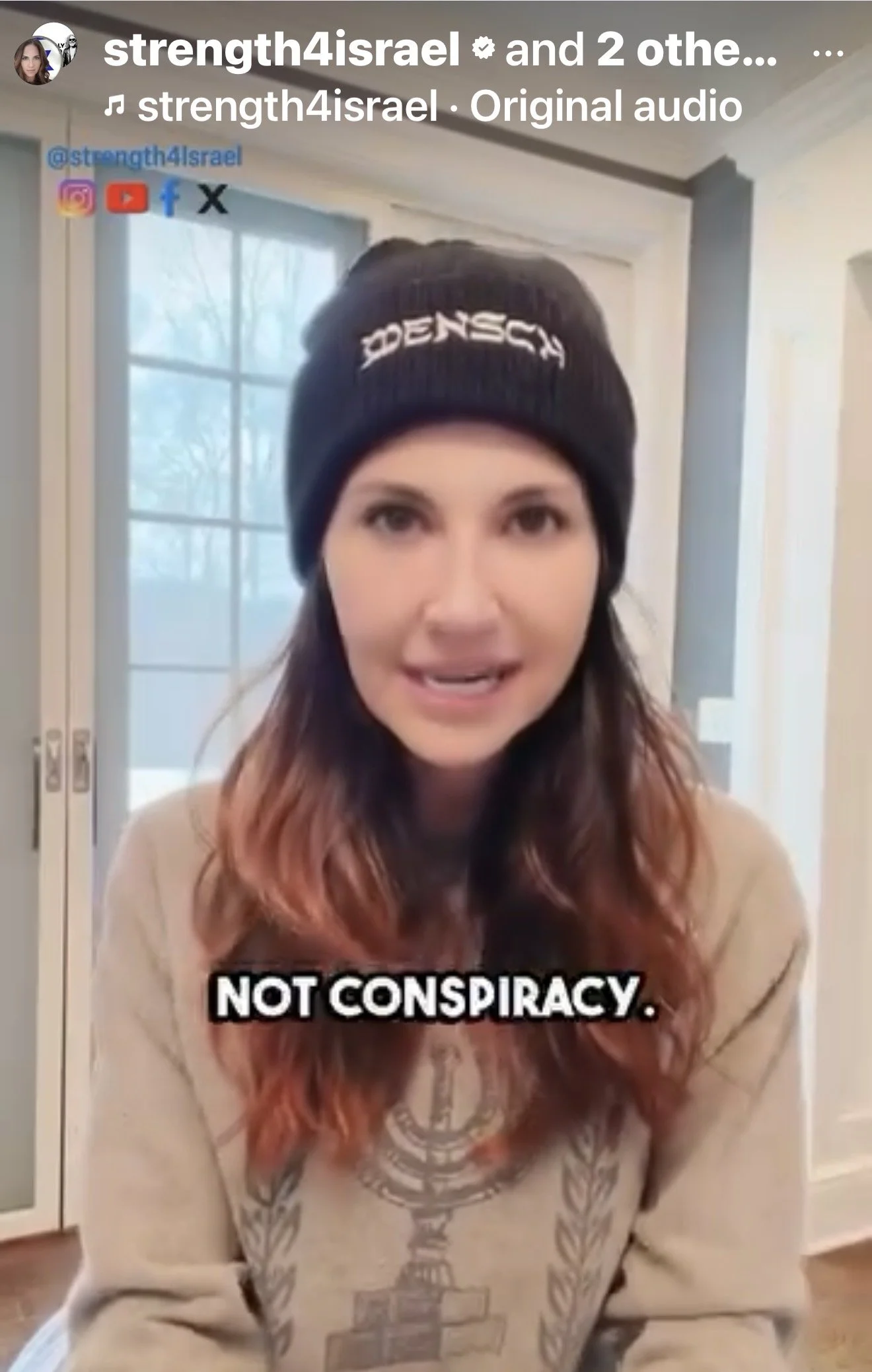 A woman with long wavy hair colored pink at the tips, wearing a black beanie with white text, a beige sweater with a design, sitting indoors with a large window and closed blinds in the background.
