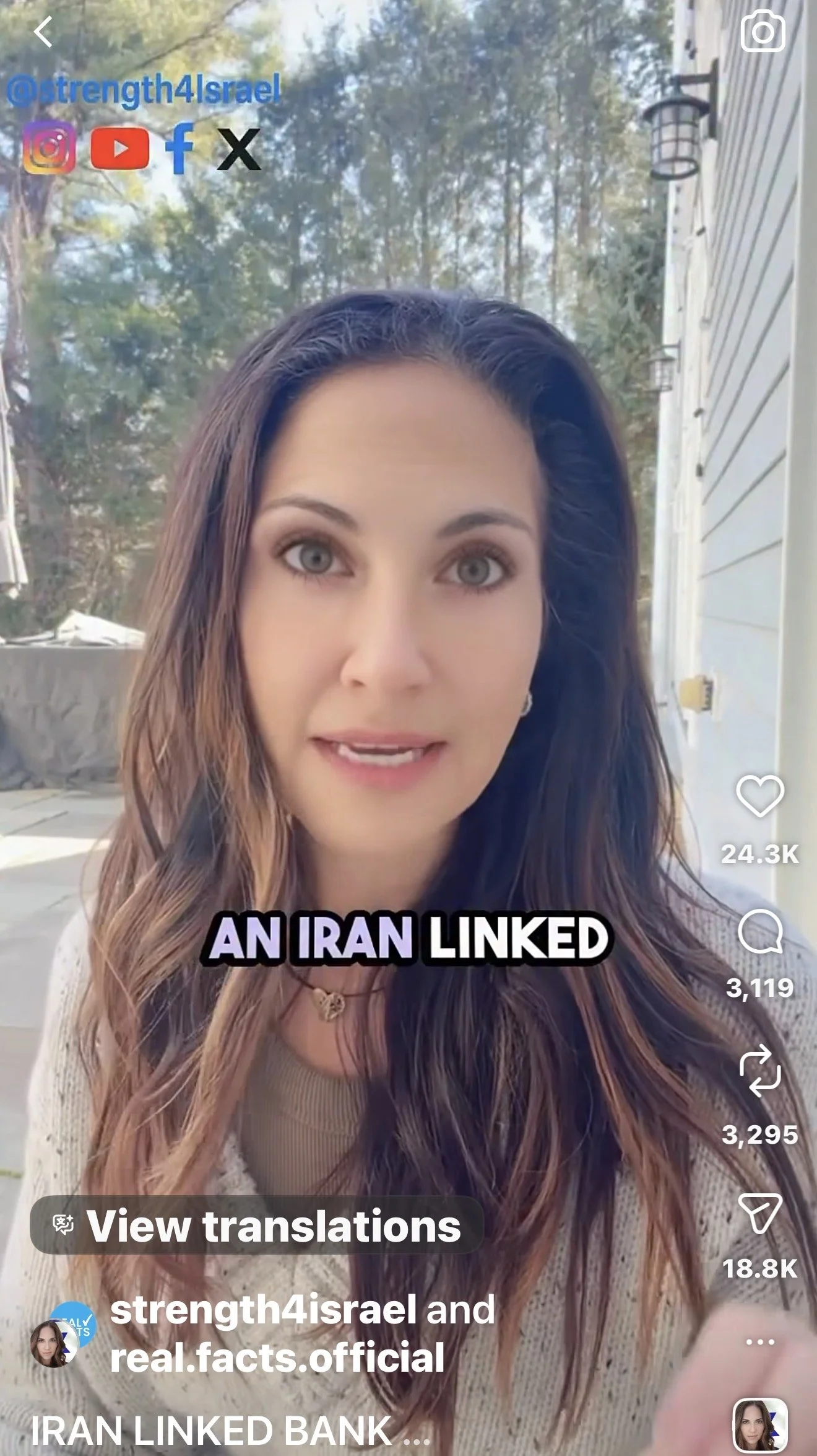 A young woman with long wavy brown hair with blonde highlights, light skin, and gray eyes, looking at the camera, standing outdoors near a house with white siding and black light fixtures, with trees and a clear sky in the background. She is wearing a beige top and a necklace, and text overlay says "AN IRAN LINKED".