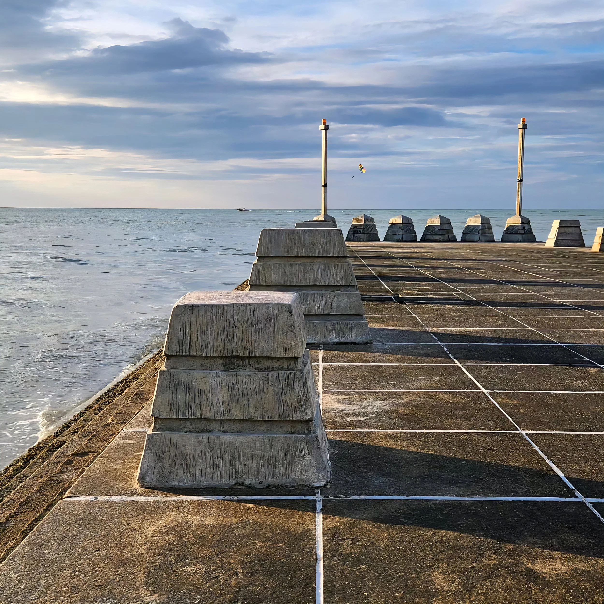 View of a pier or dock with two poles, a line connecting them, and a colorful kite flying in the sky over the water, with clouds and a boat in the distance.