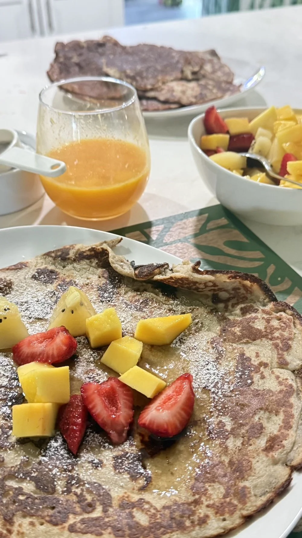 Breakfast spread including a crepe topped with strawberries, mango, and powdered sugar, a bowl of fruit salad with strawberries, mango, and banana, a glass of orange juice, and a plate of cooked meat in the background.
