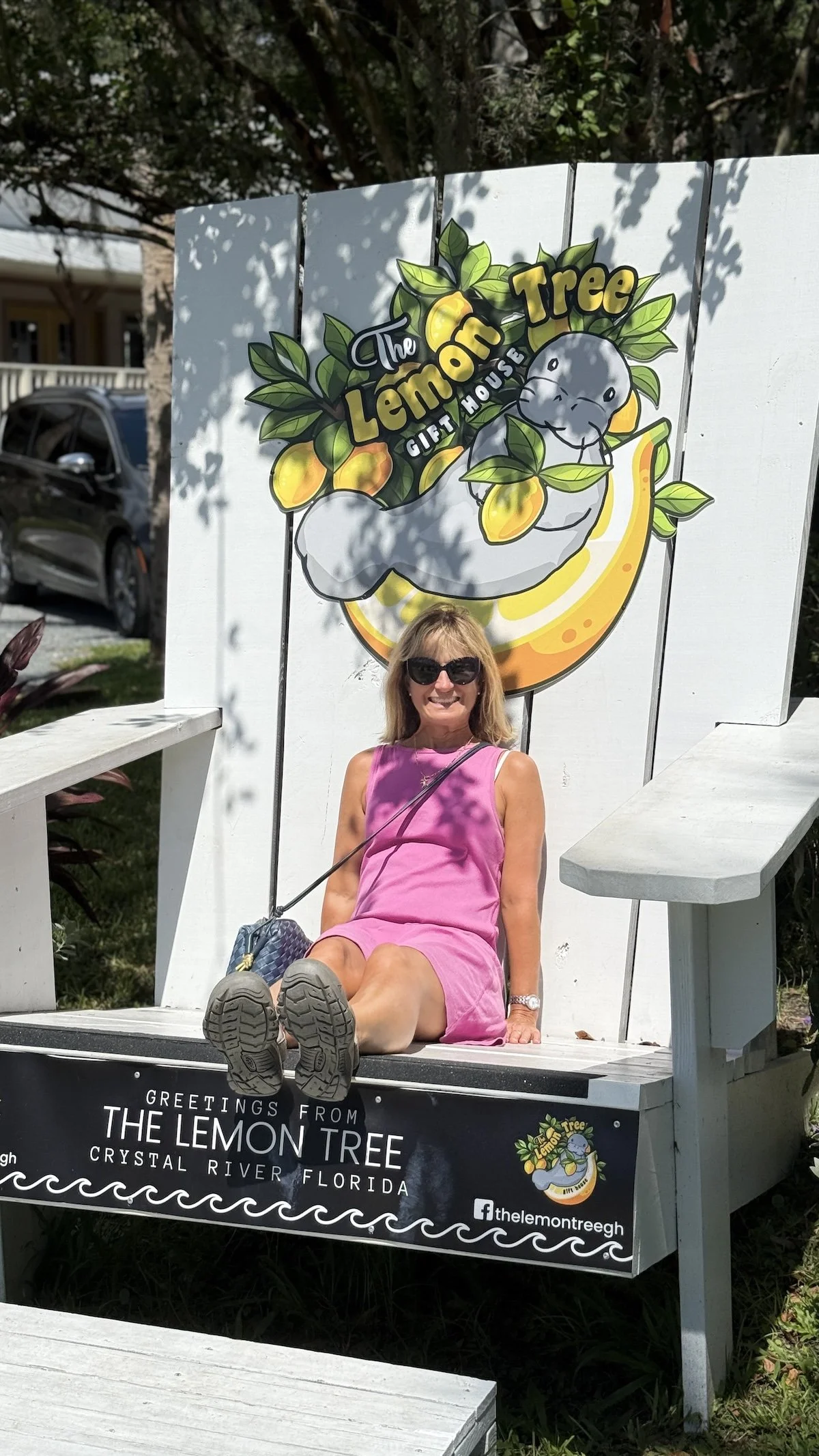 A woman wearing sunglasses and a pink dress sitting on a large decorative chair shaped like an lemon slice in front of a sign for The Lemon Tree gift house in Crystal River, Florida.
