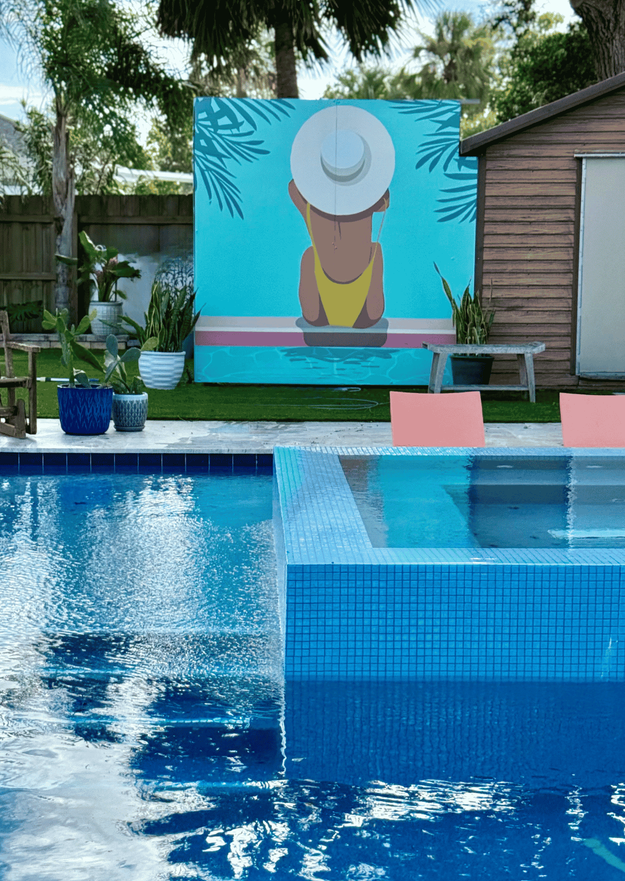 A swimming pool with clear blue water and tile edges, surrounded by a backyard with green grass, potted plants, outdoor furniture, and a wooden shed. There's a large mural or painting of a woman in a yellow swimsuit with a sunhat on the wall.