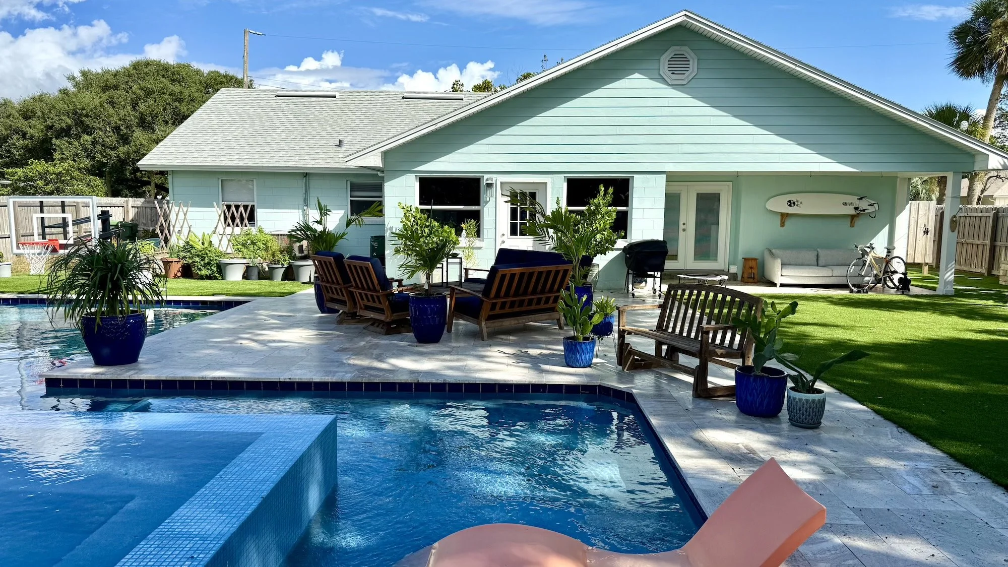 A backyard with a swimming pool, surrounded by potted plants and outdoor furniture, with a house in the background. The house is painted light blue and has a covered patio area with a sofa, a bicycle, and a surfboard mounted on the wall.