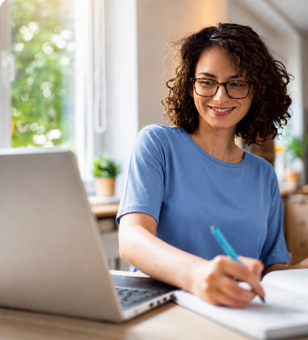 Woman with curly hair and glasses writing in a notebook at a desk with a laptop